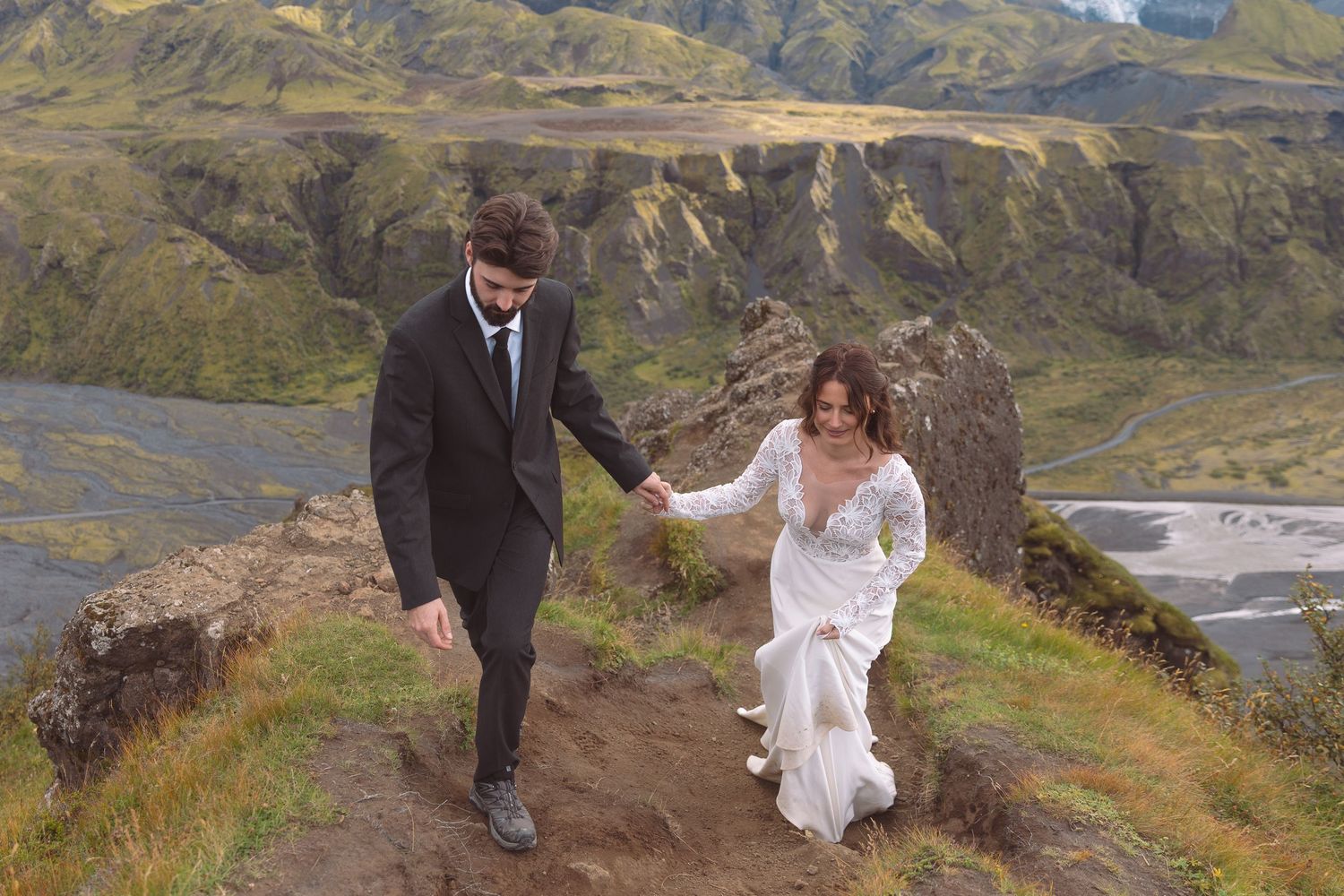 Bride and groom walk hand in hand along a scenic Iceland mountain trail in wedding attire.