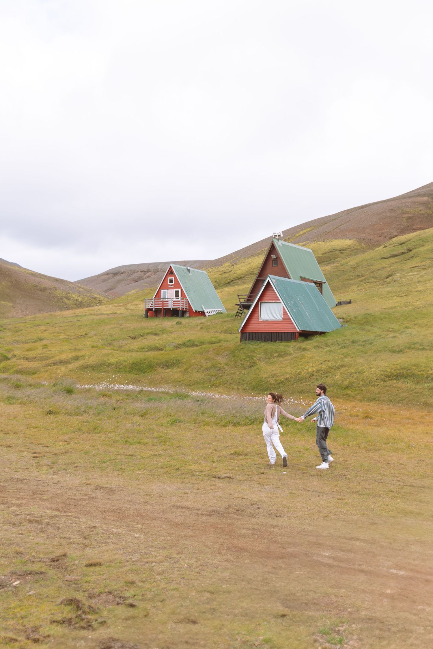 Romantic couple holding hands walking toward red and green A-frame cabins on a scenic Icelandic hillside meadow.