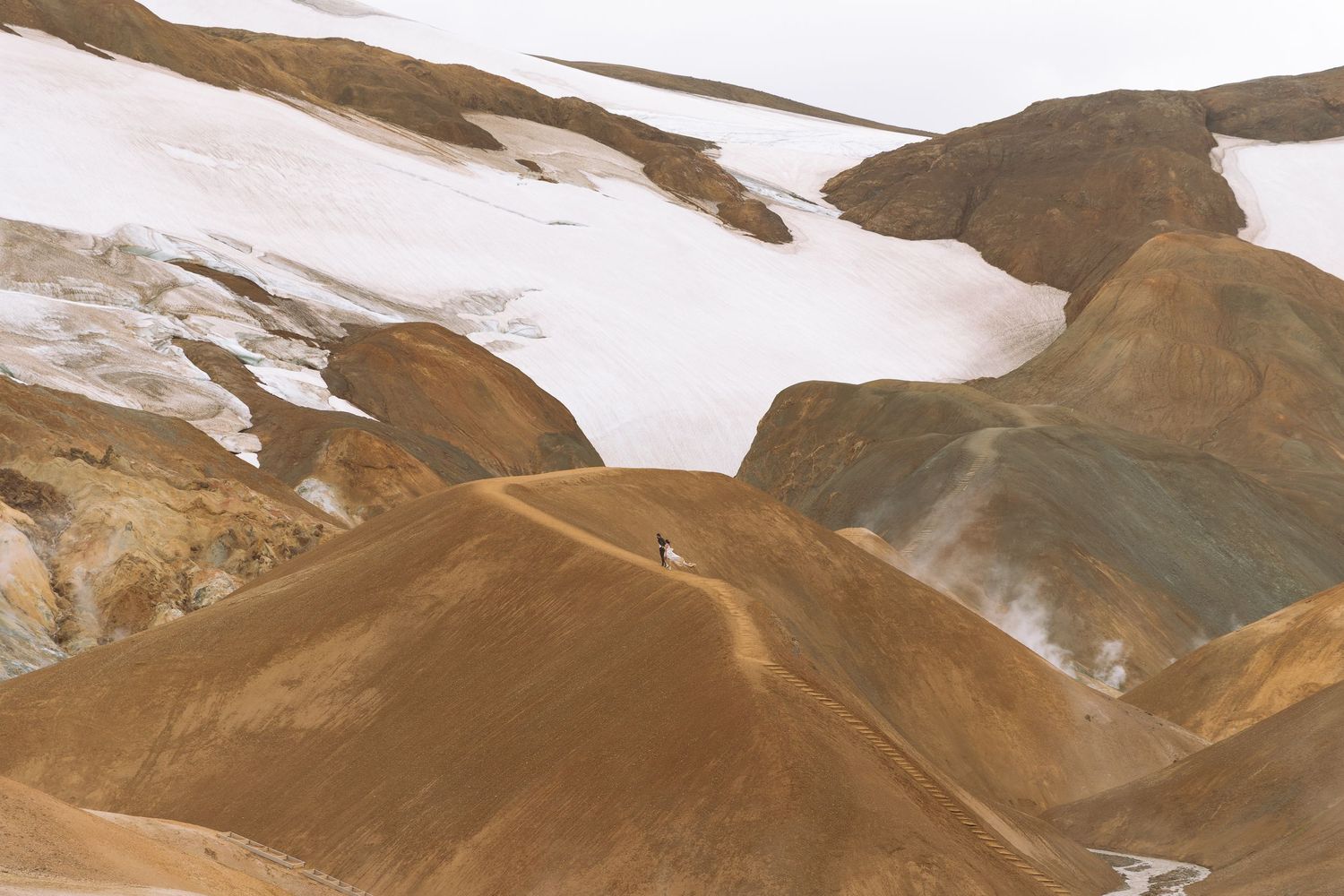 Tiny figure standing atop vibrant rhyolite hills surrounded by snow and geothermal steam in Iceland.