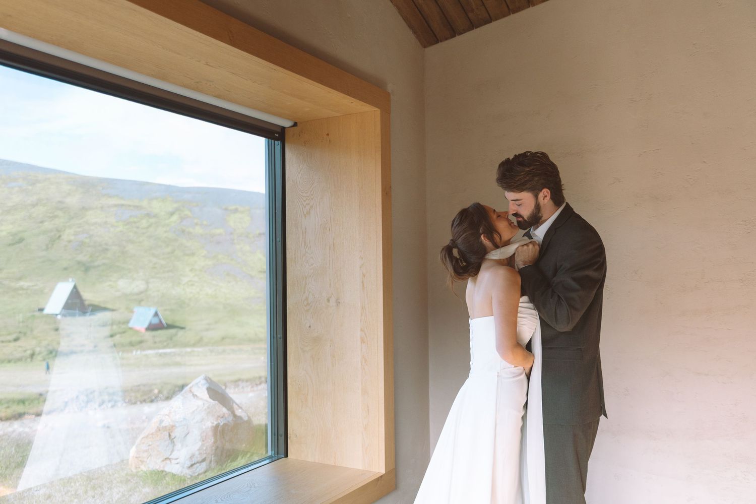 Newlyweds sharing a tender kiss near a large window overlooking a lush green landscape.