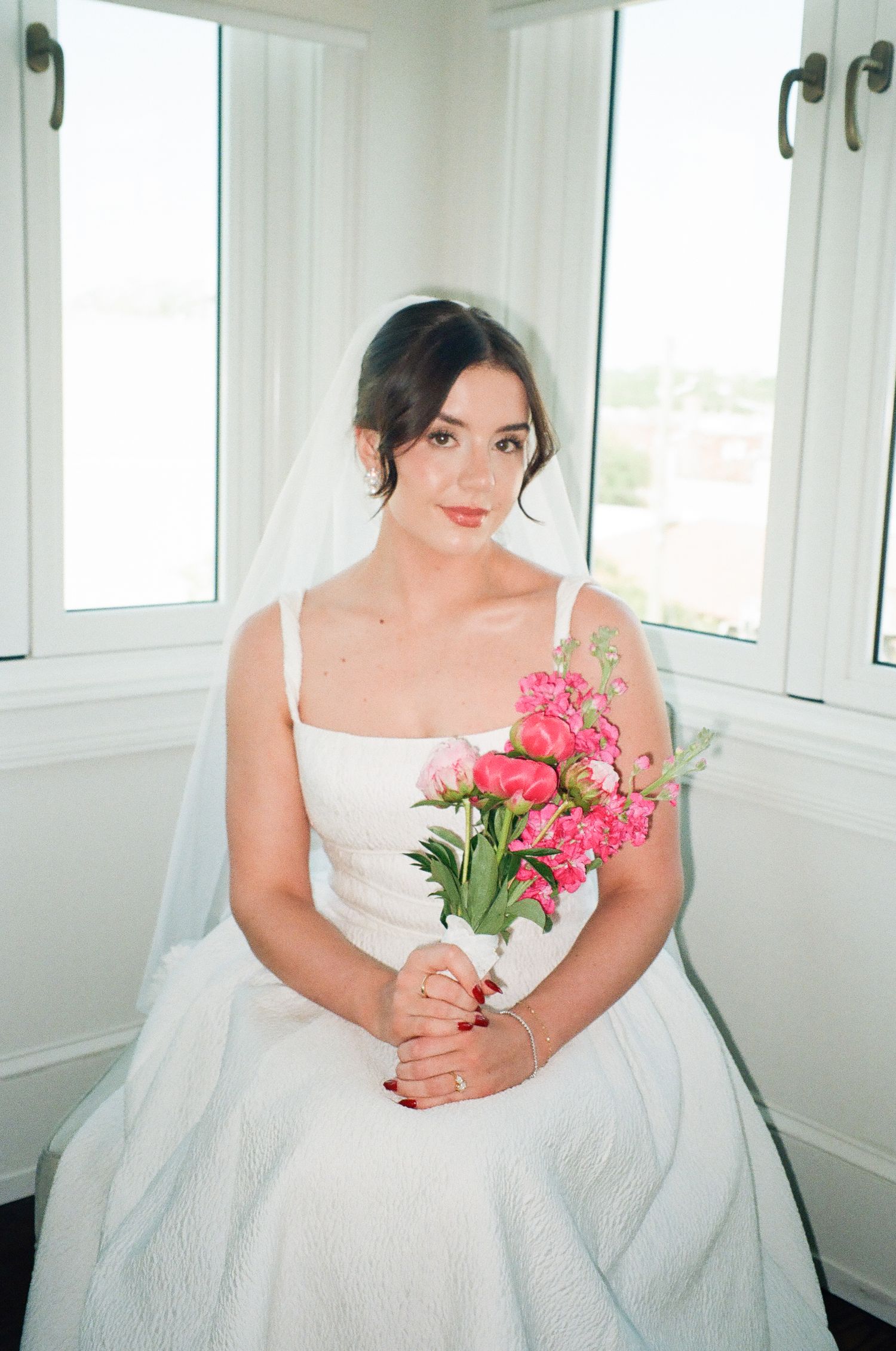 Elegant bride in a white gown and veil sits by a bright window holding a vibrant pink floral bouquet.