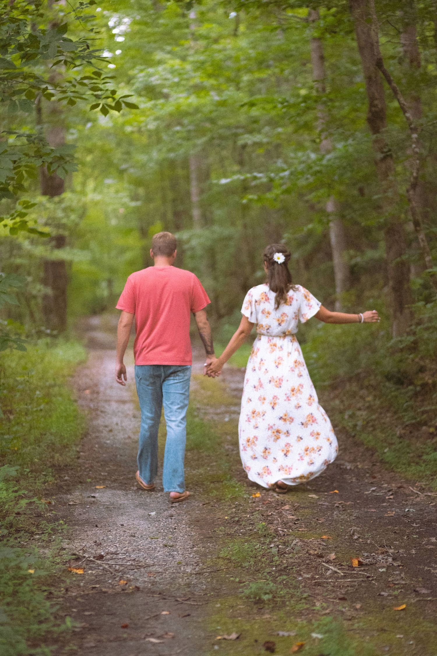 Couple walking hand in hand down a lush green forest path from behind.