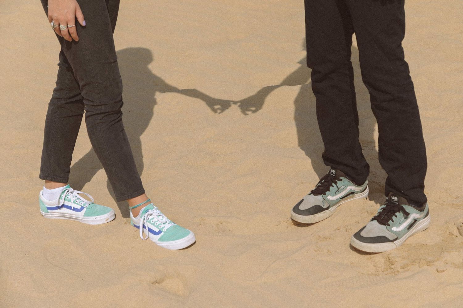 Close-up of couple holding hands on sand, wearing colorful sneakers with heart-shaped shadow.
