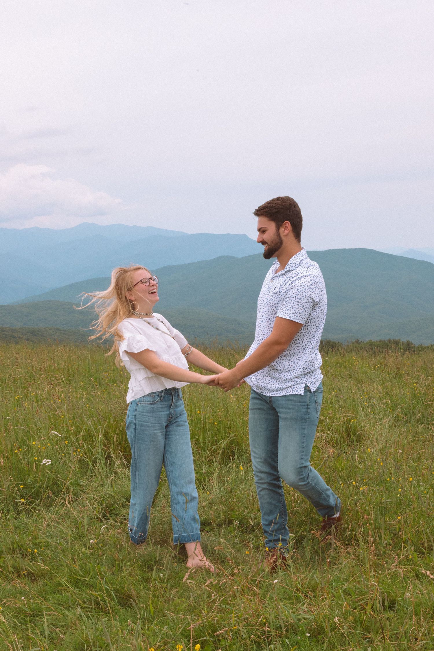 Joyful couple holding hands and dancing in a green mountain meadow, woman laughing with hair blowing.
