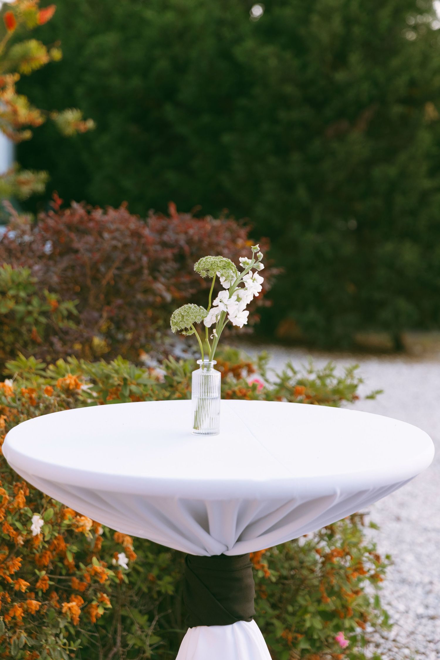 Minimalist cocktail table with white linen and small floral arrangement outdoors in garden setting.