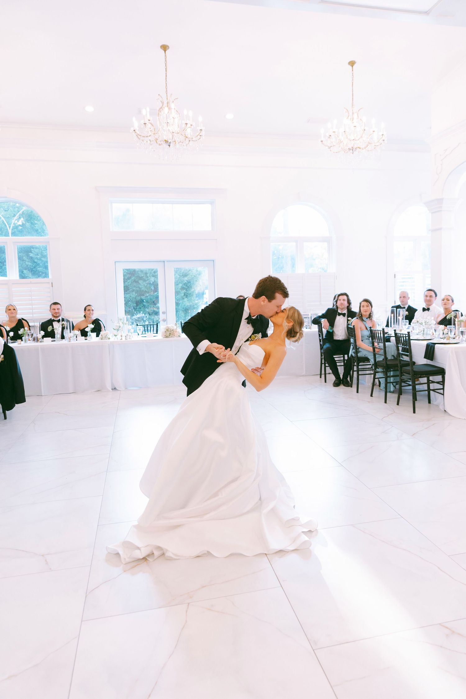Bride and groom share romantic dip kiss during first dance in elegant white ballroom.