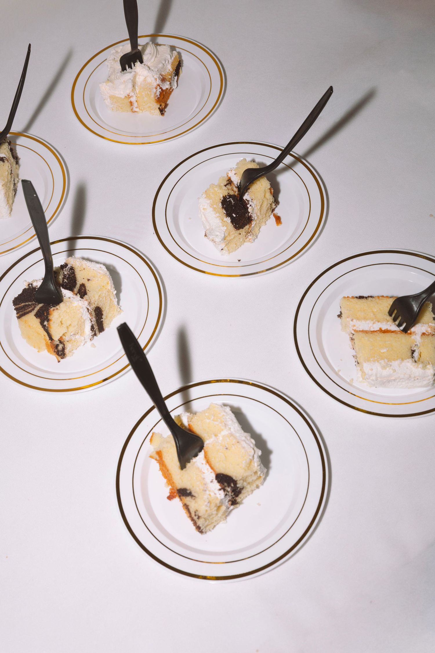 Six gold-rimmed plates each holding a slice of layered cake with white frosting and chocolate, served with black forks.