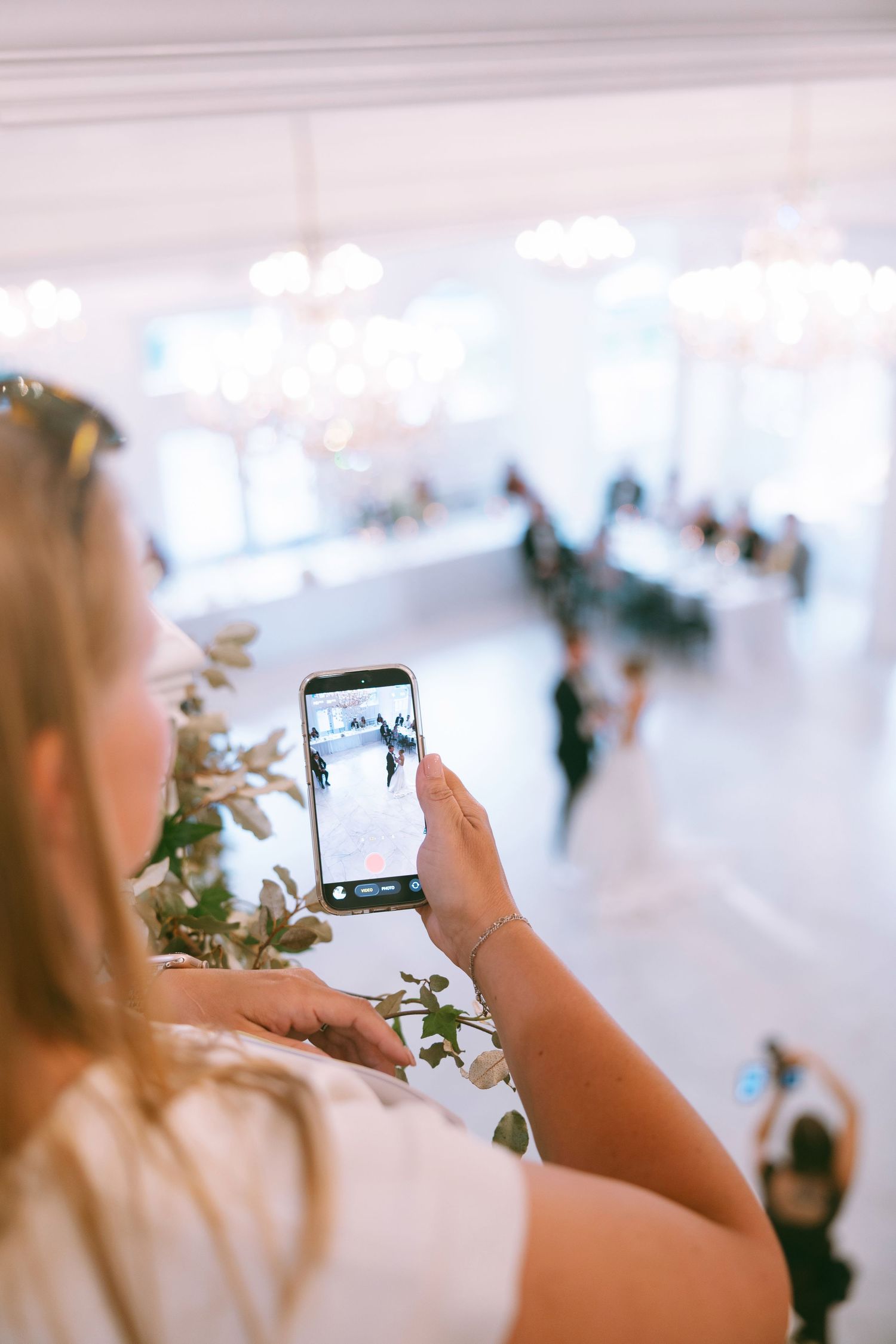 Woman photographing wedding reception dancers on smartphone in elegant white ballroom with chandeliers.