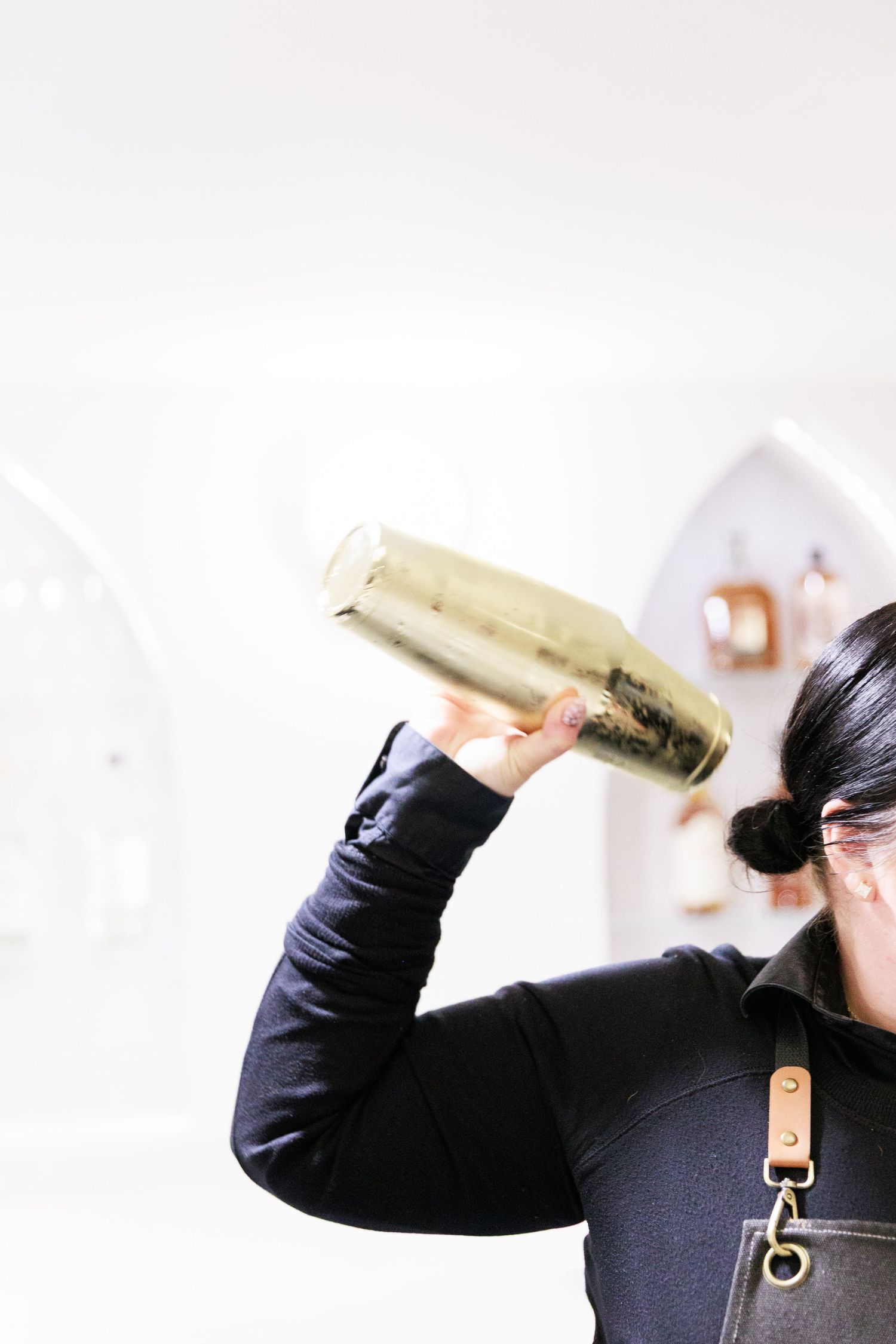 Bartender in black apron shaking a gold cocktail shaker in a modern bar setting.