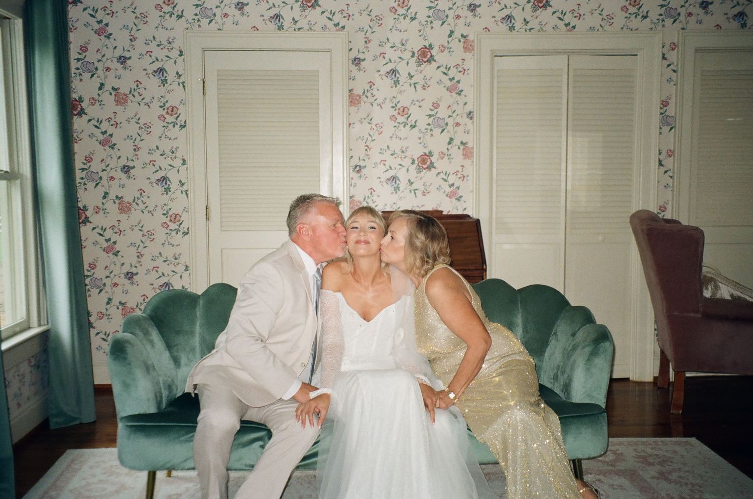 Bride in white gown receives kisses on cheeks from parents on teal velvet sofa in floral wallpapered room.