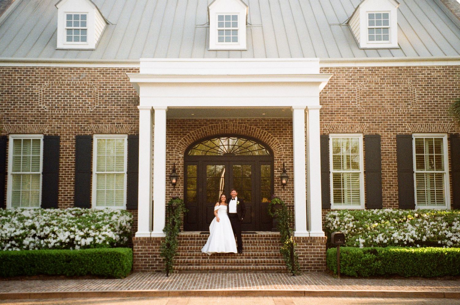 Bride and groom pose at entrance of elegant brick colonial wedding venue with white columns and manicured hedges.