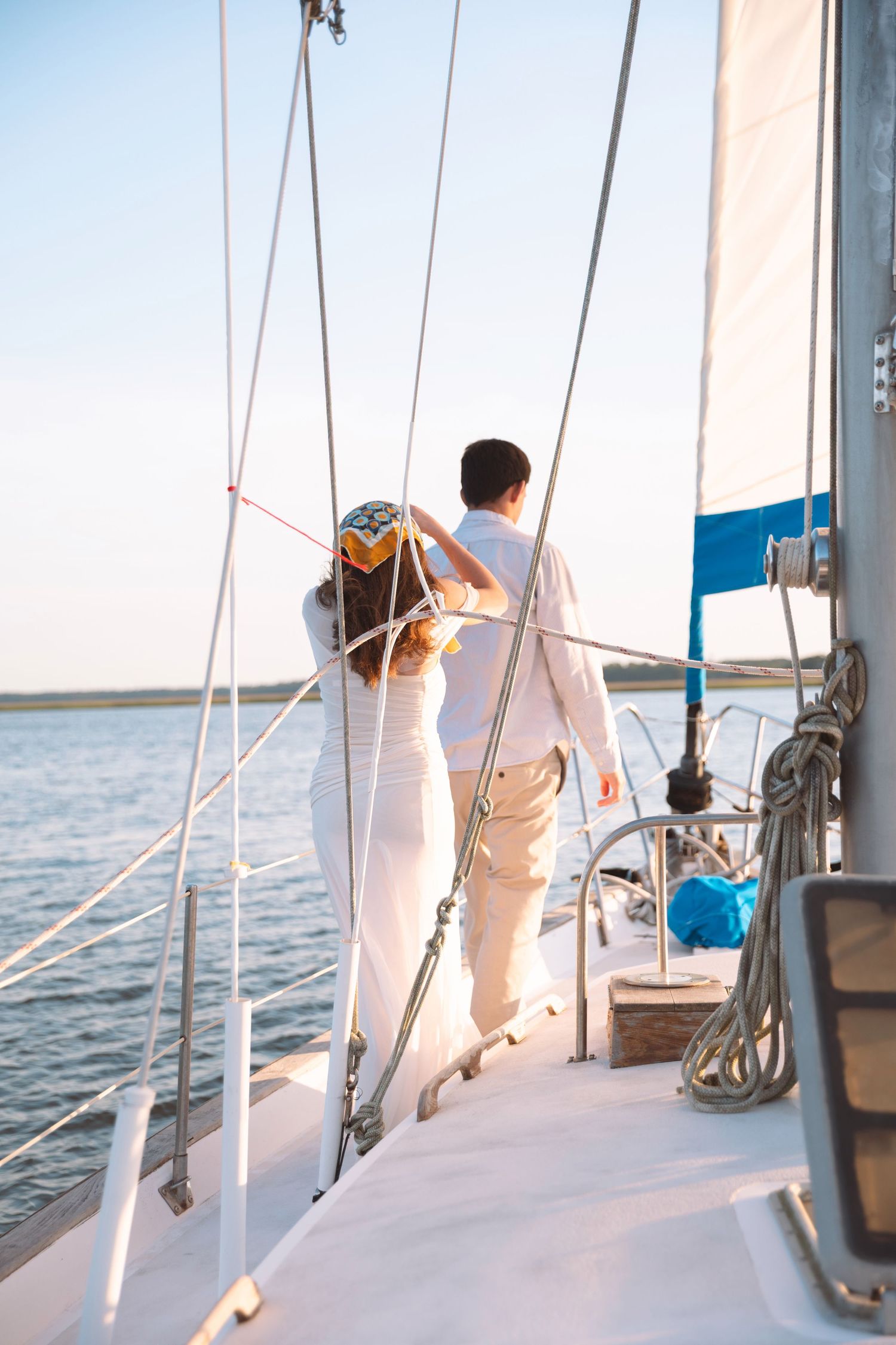 A couple dressed in white stands together on the bow of a sailboat, gazing at the calm open water.