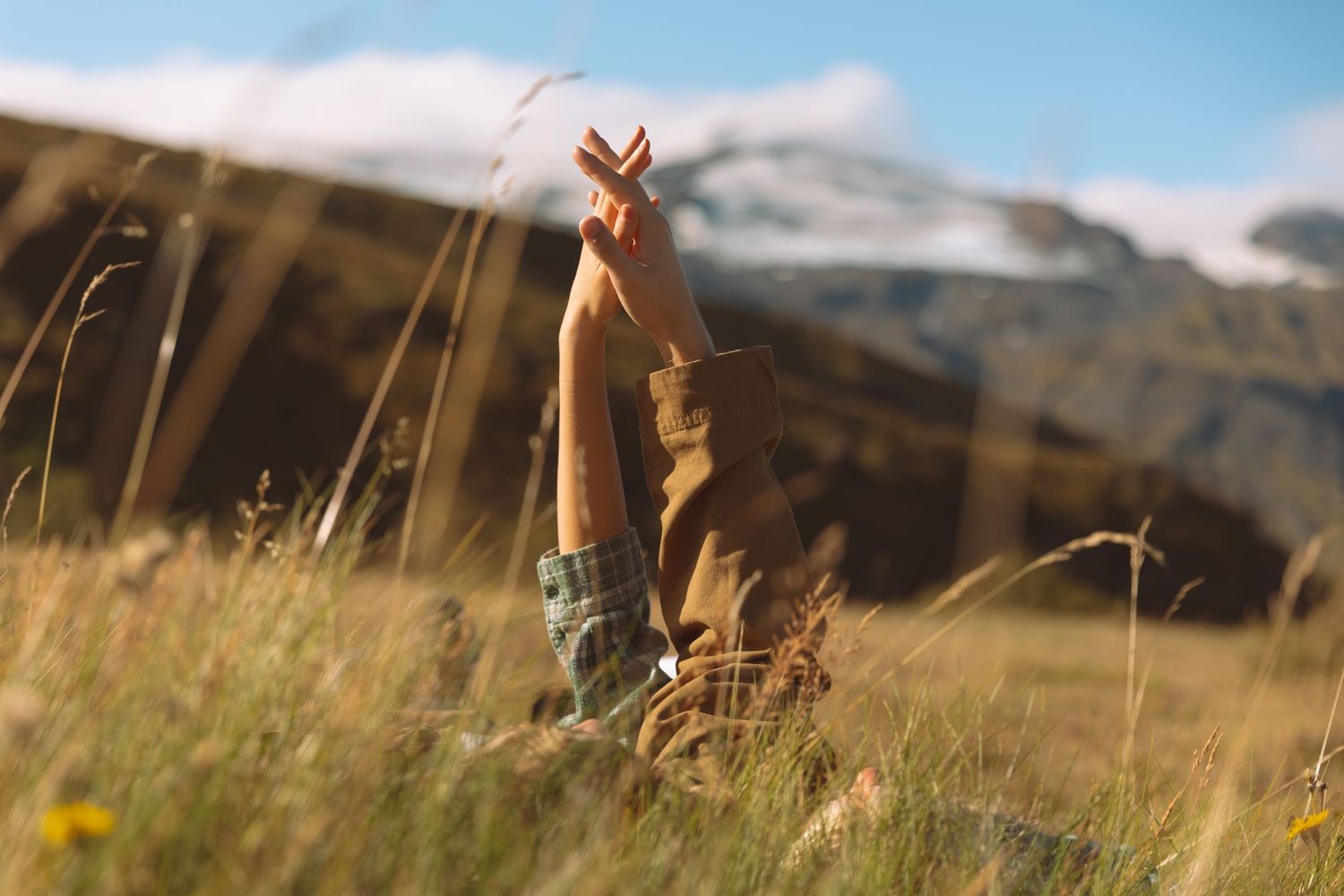 Person lying in tall grass with hands raised toward mountains and blue sky in the background.