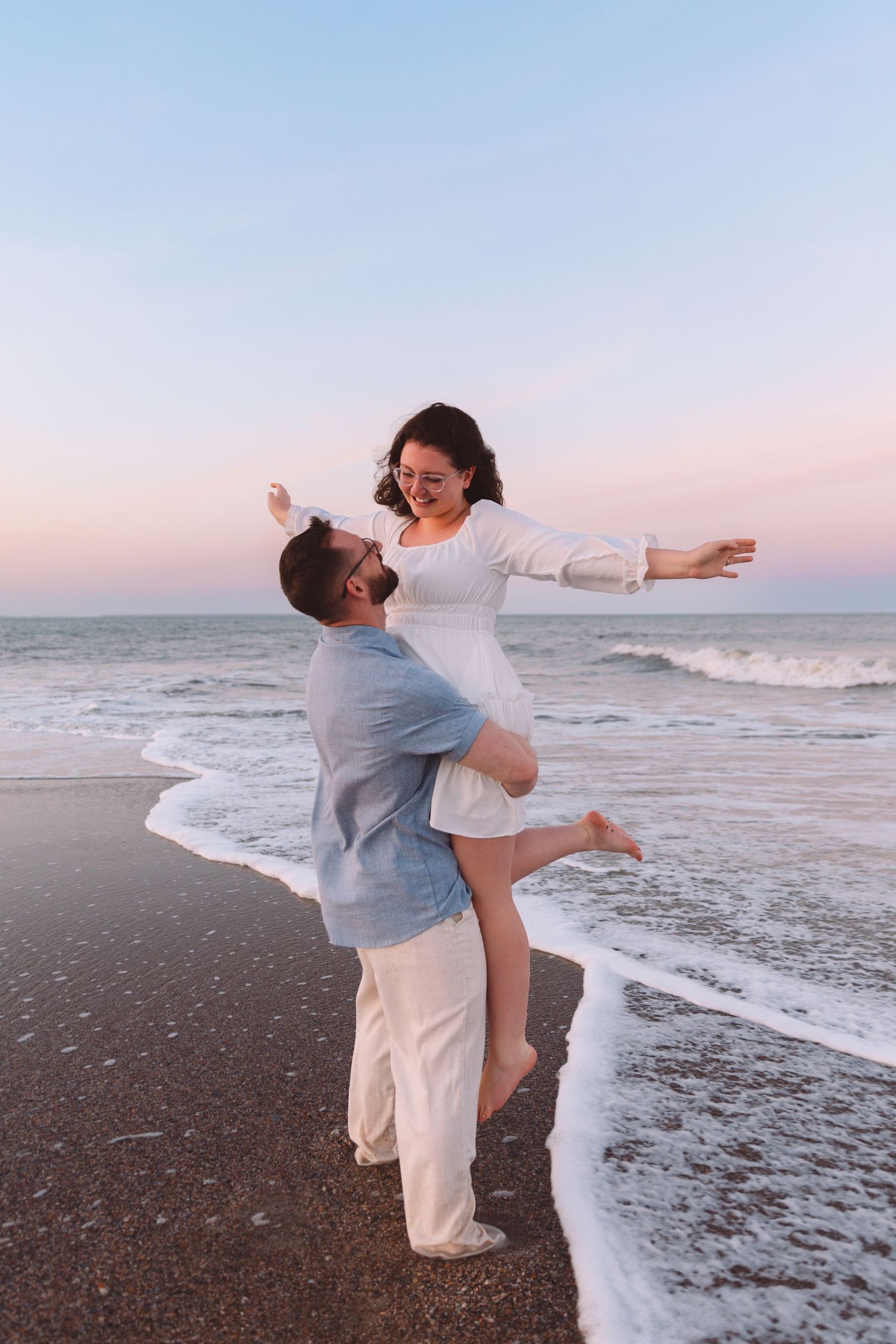 Happy couple at beach sunset, man lifting woman in white dress as waves wash ashore in pink twilight.