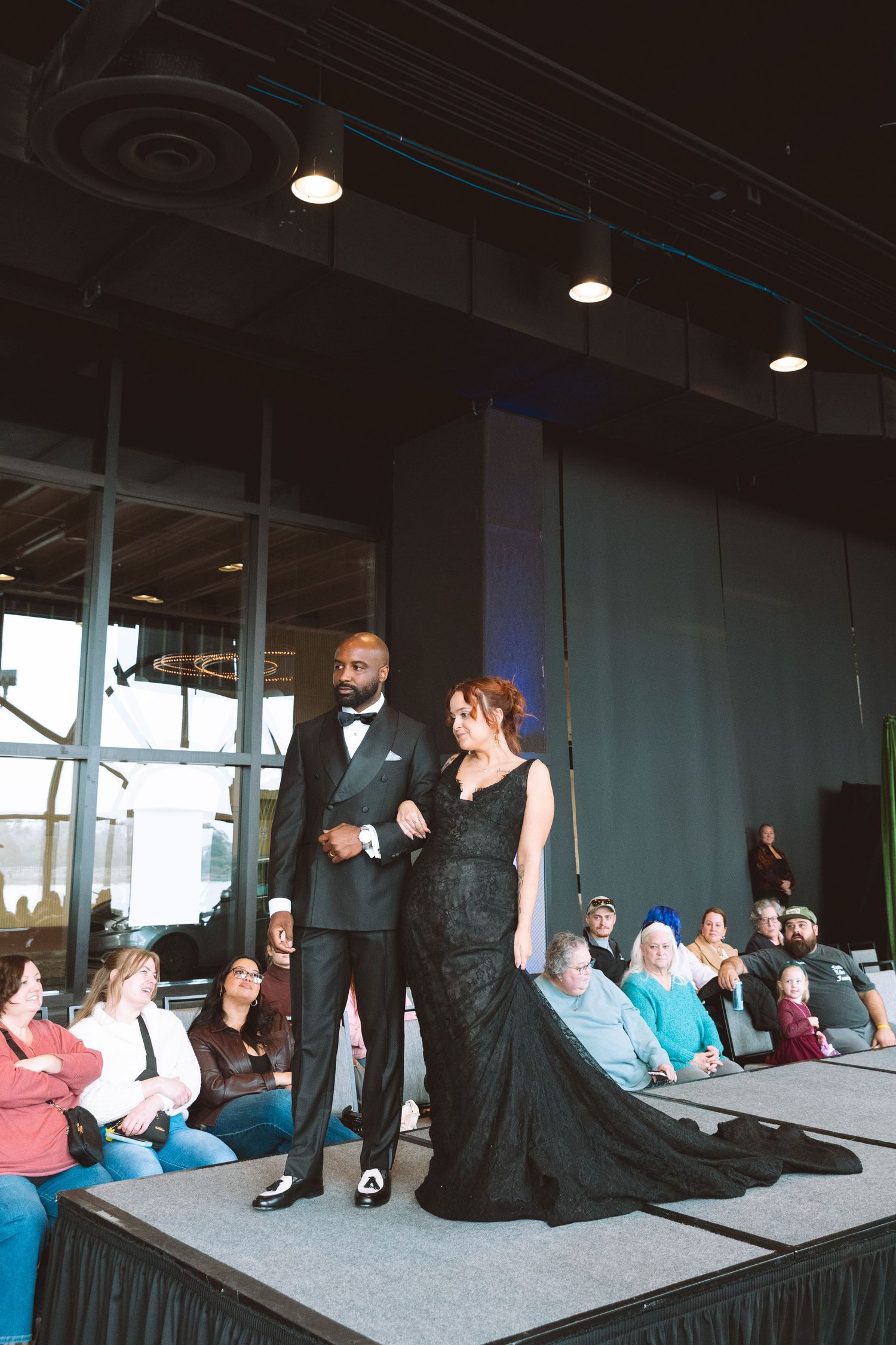 Couple in elegant black formal wear on runway at fashion show, man in tuxedo and woman in black gown.