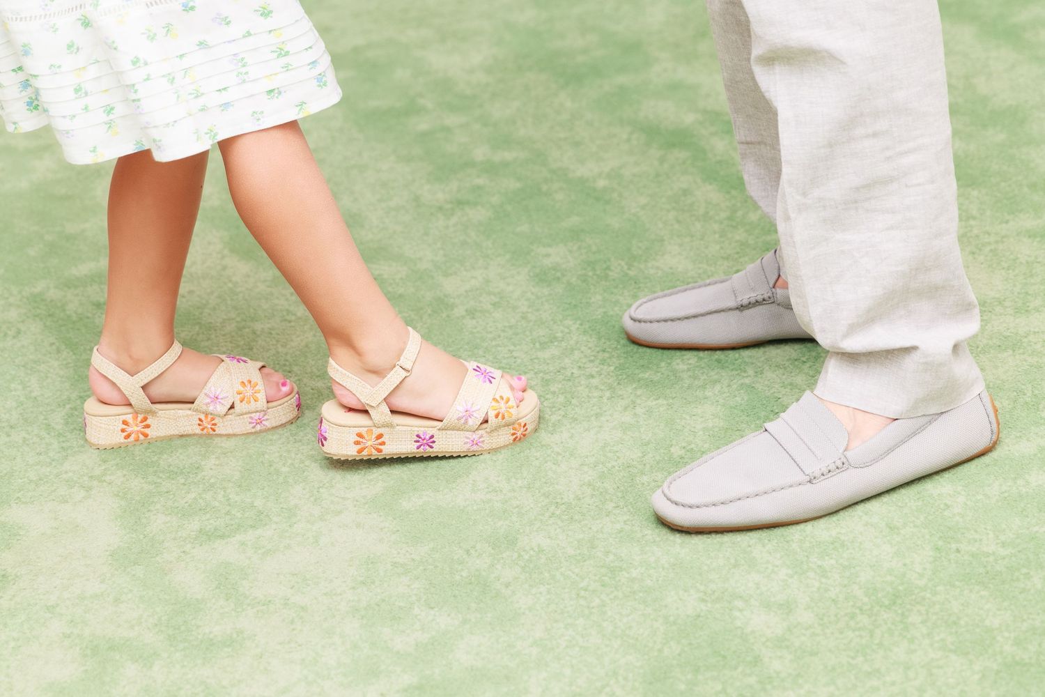 Close-up of a child in floral sandals and an adult in gray loafers standing on green grass.
