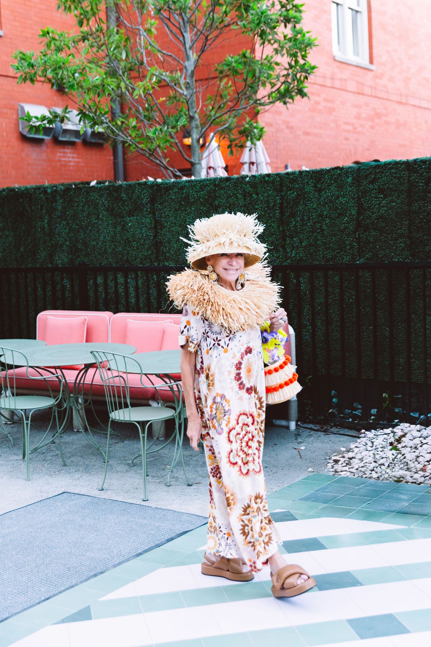Woman in floral maxi dress, straw hat, and raffia necklace poses in a stylish outdoor patio with green hedges.