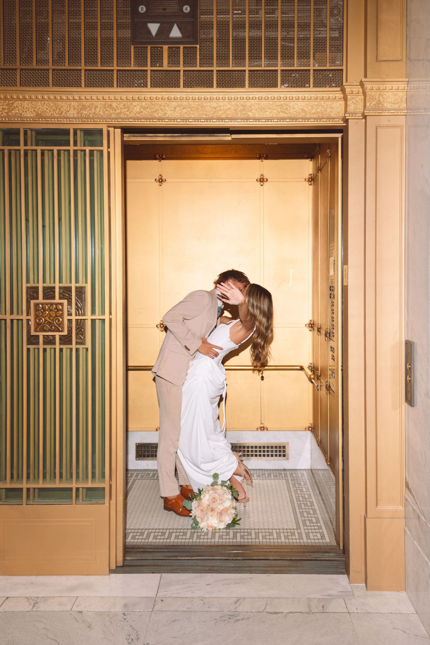 Newlyweds sharing a romantic kiss inside a ornate gold elevator during their wedding day.