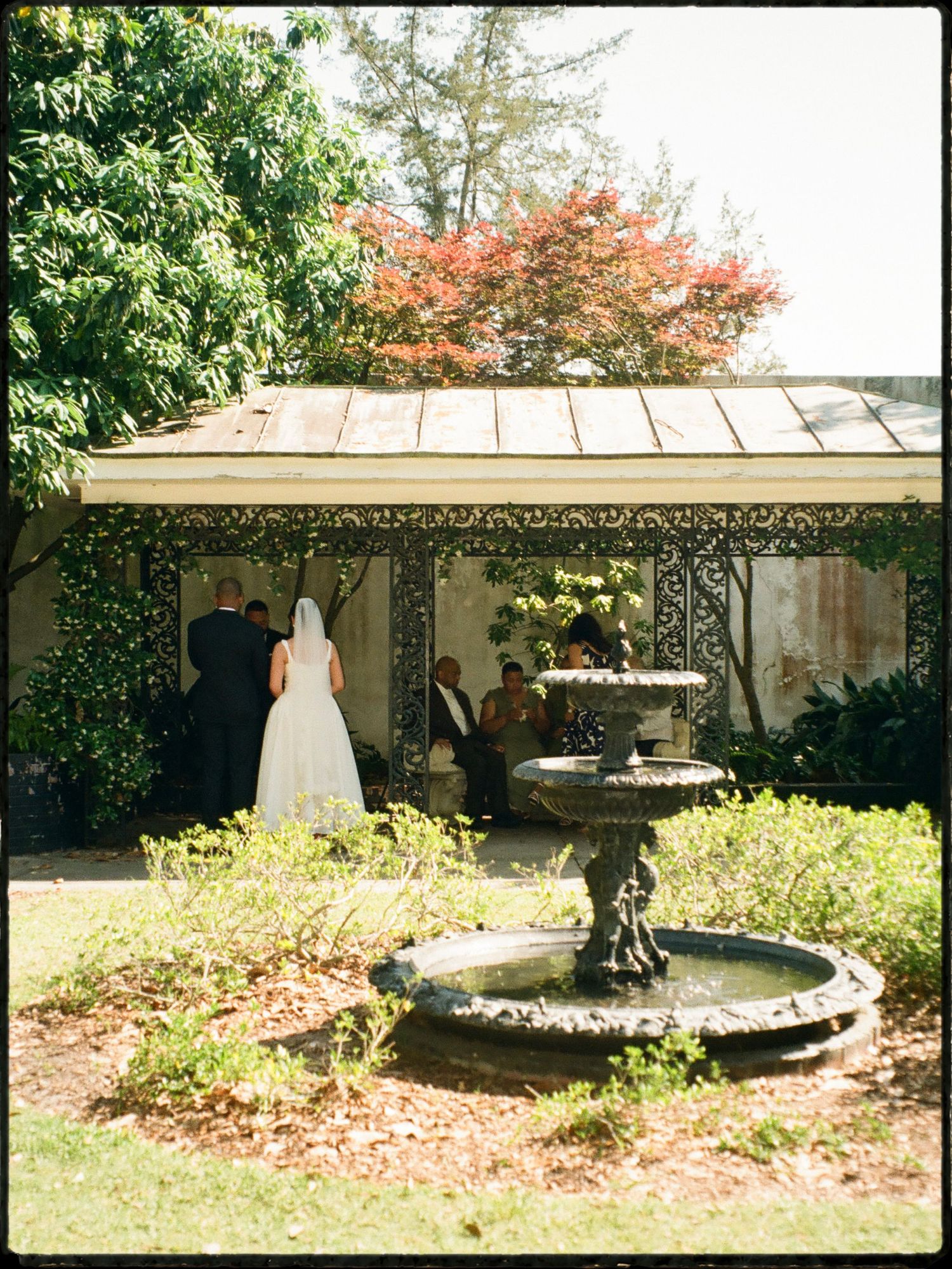 Bride in white gown and veil standing near a stone fountain in a sunlit garden venue.