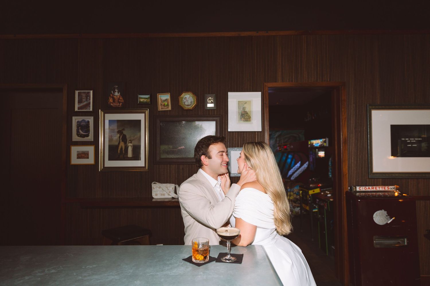 Bride and groom share a romantic moment at a vintage bar adorned with framed photos and warm lighting.