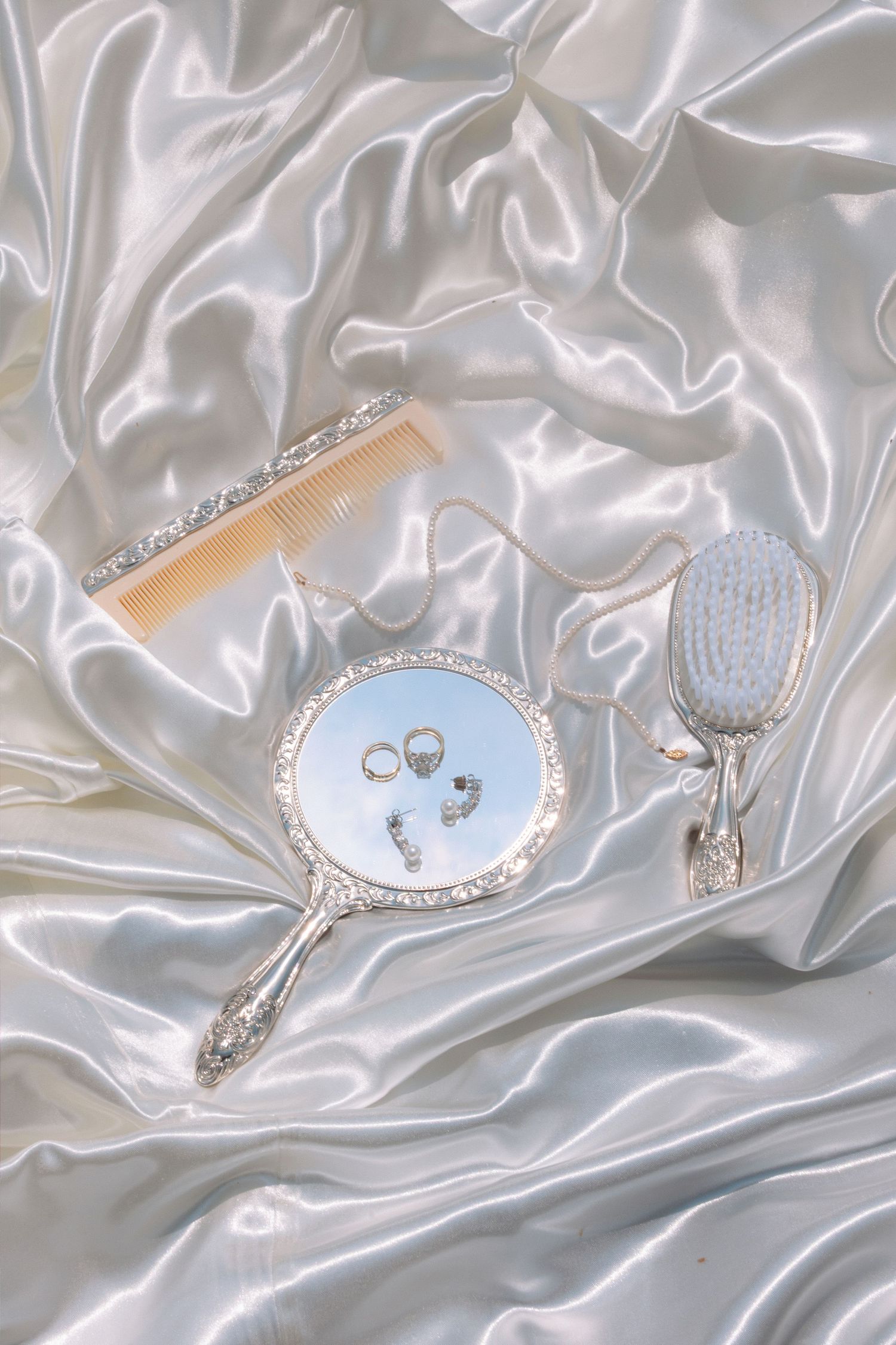 Elegant silver vanity set with hand mirror, brush, and comb with rings on white satin fabric.