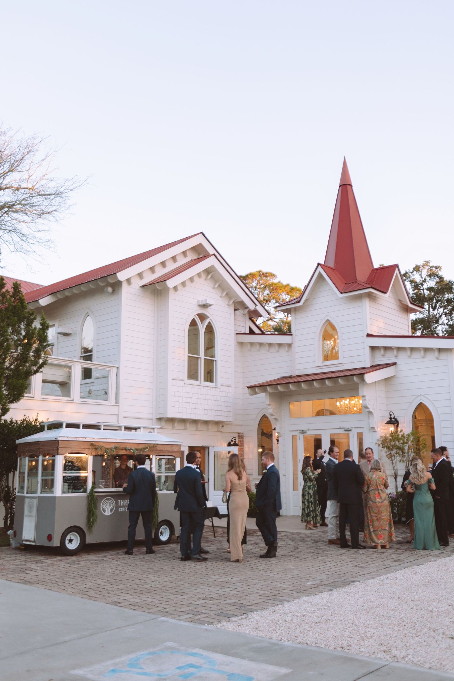 Guests mingling outside white chapel wedding venue with red steeple and vintage food truck at dusk.