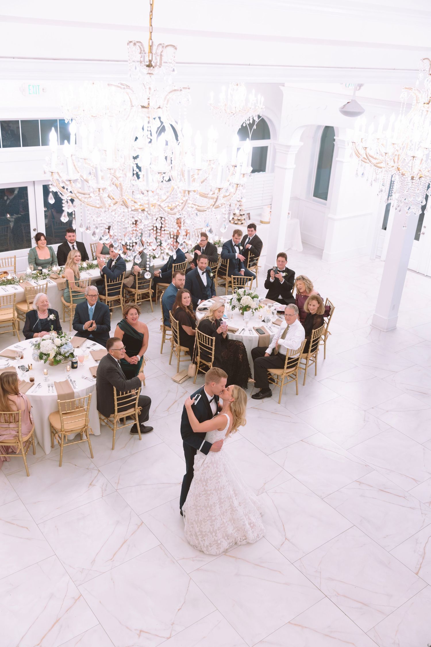 Newlyweds share their first dance in an elegant white ballroom with crystal chandeliers and guests.