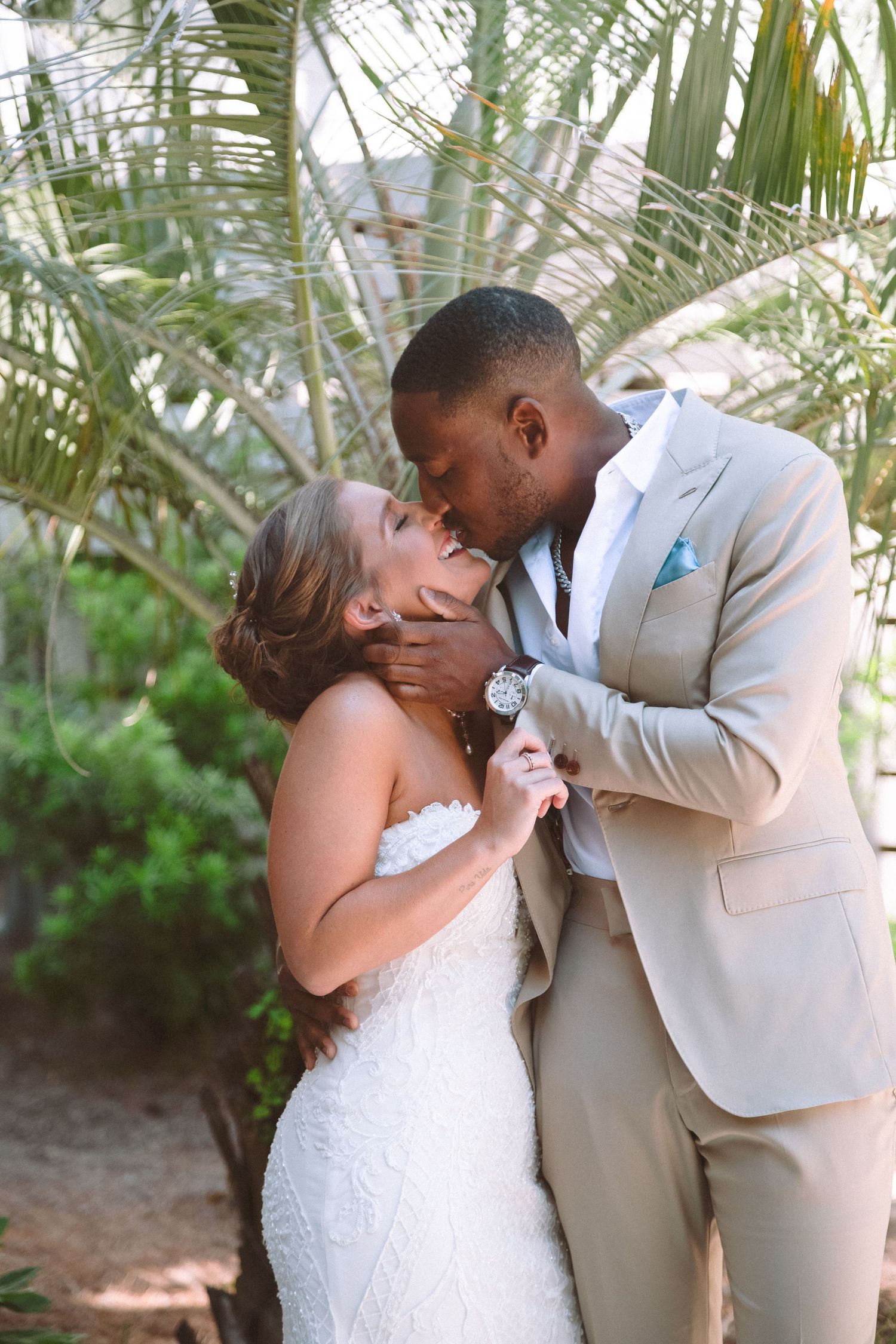 Groom tenderly kisses bride's forehead during romantic outdoor first look moment beneath lush palm trees.