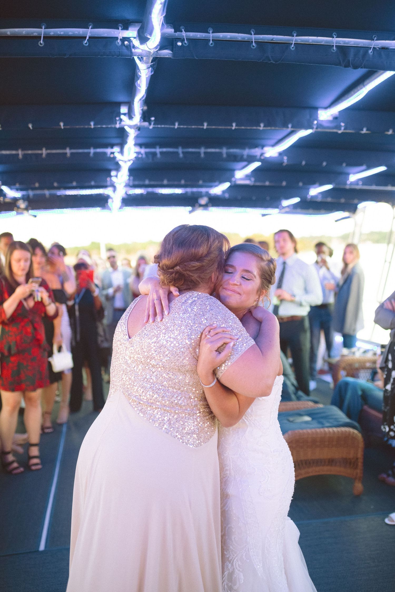 Bride embraces a guest in a gold sequin dress surrounded by wedding reception crowd.