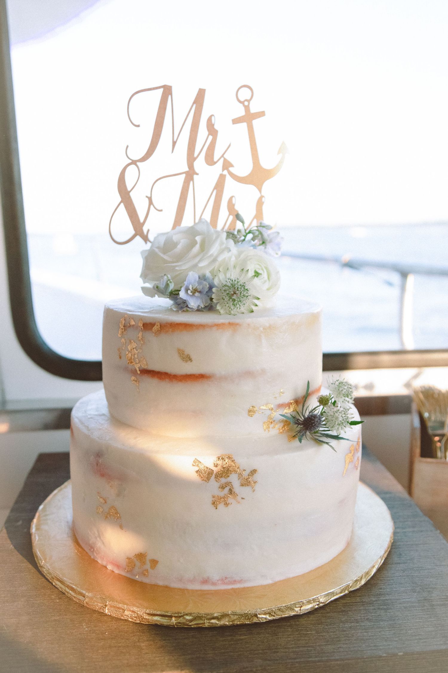 Two-tier white and gold wedding cake with Mr & Mrs anchor topper and white floral decorations.