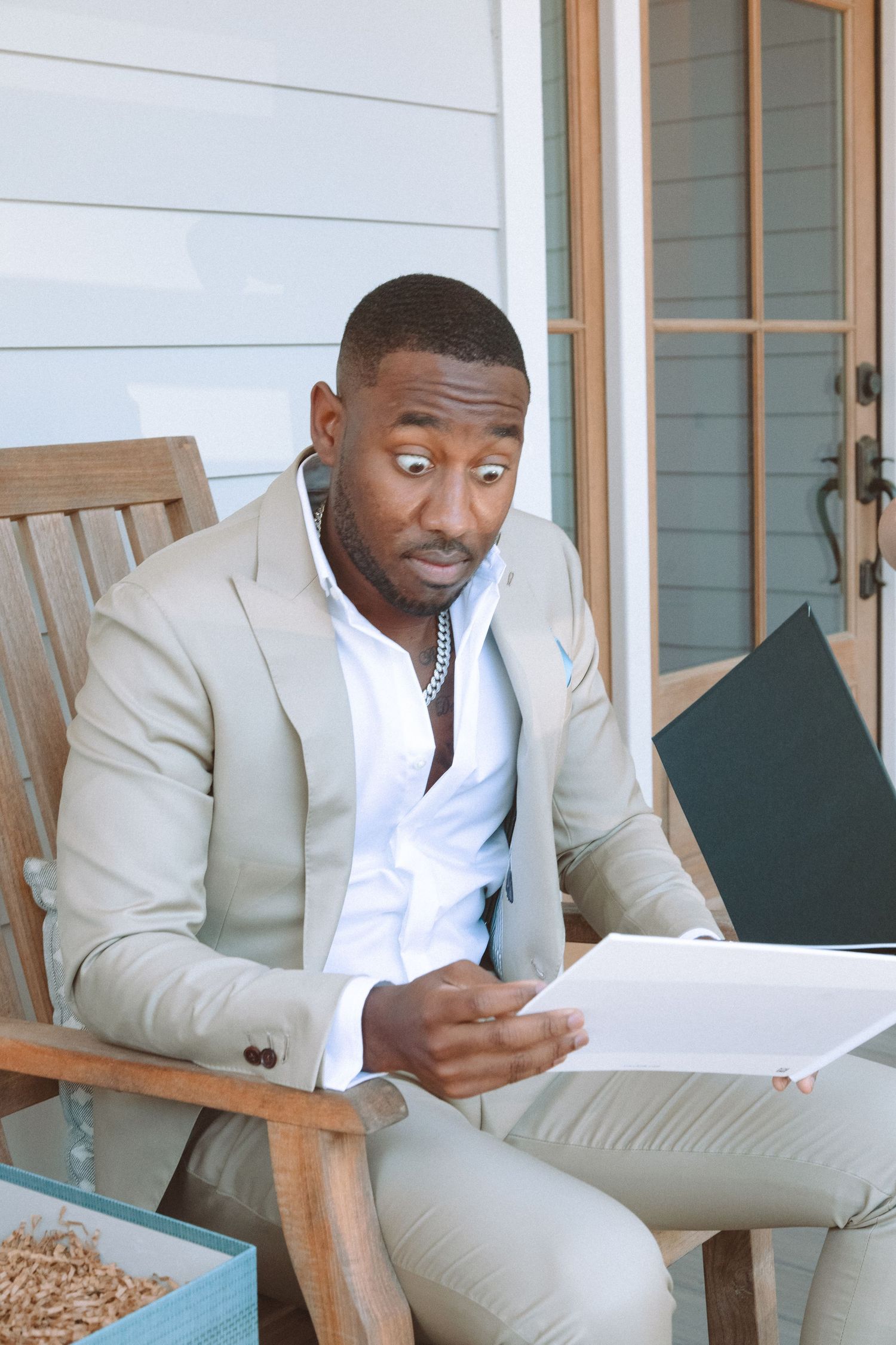Elegant man in beige suit reading documents with surprised expression while seated outdoors on porch.