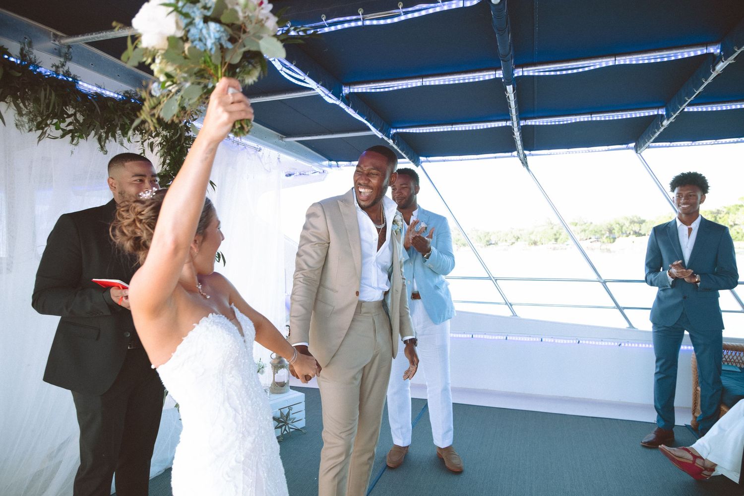 Bride raises bouquet in celebration as groom and guests cheer after wedding ceremony.