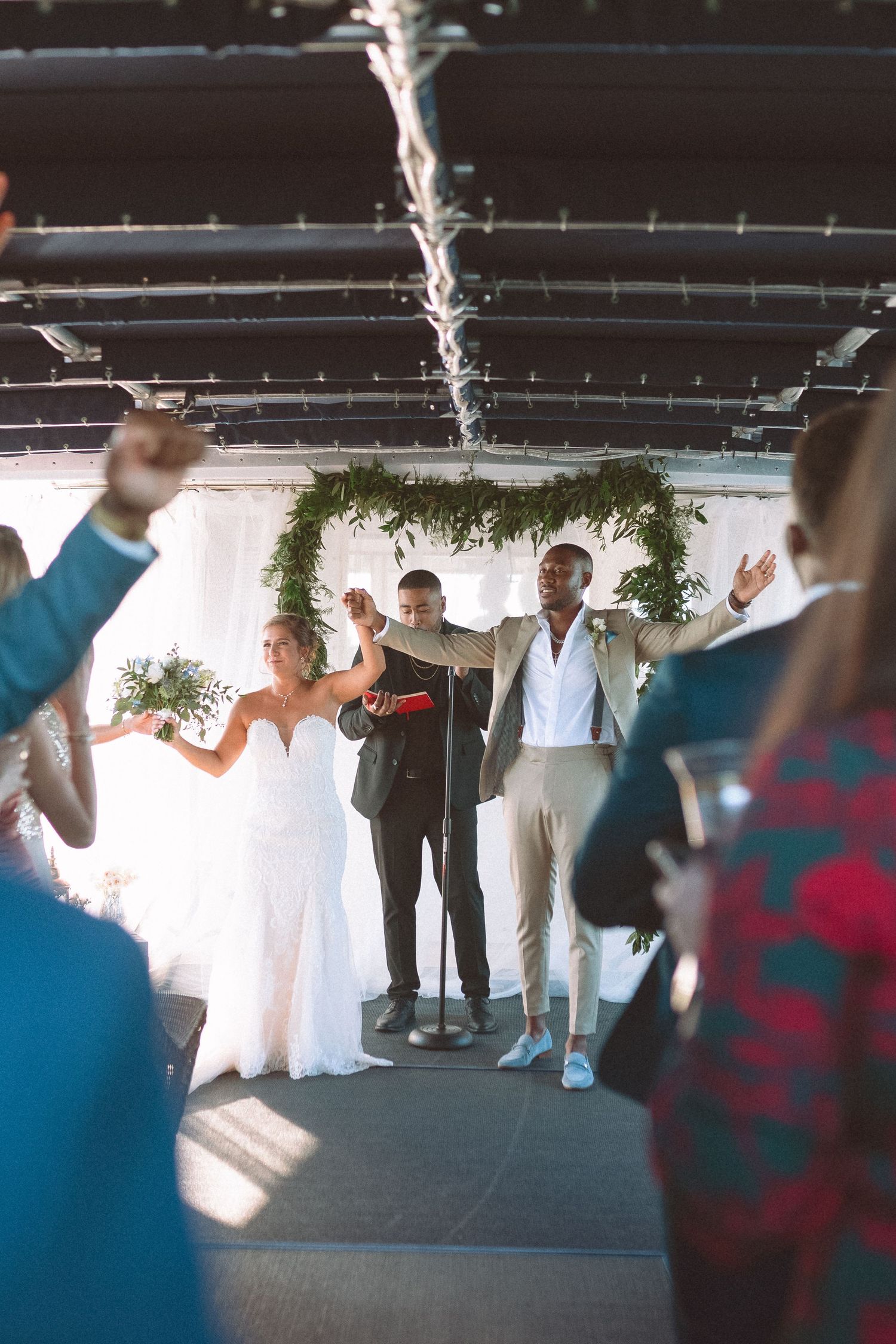 Newlyweds celebrating with raised arms under greenery arch as guests cheer at wedding.