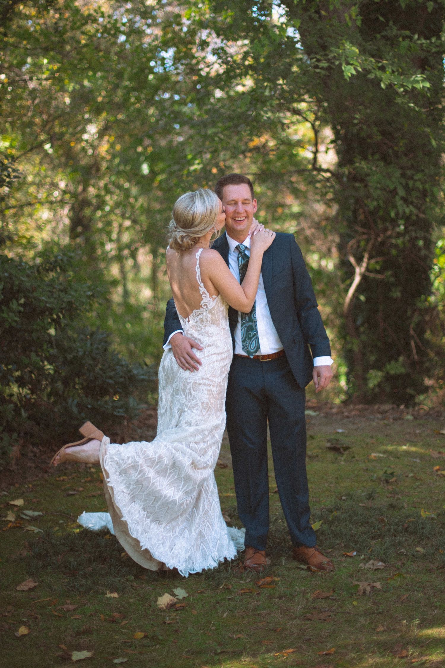 Bride in lace gown kisses groom in navy suit on cheek outdoors in lush green garden setting.
