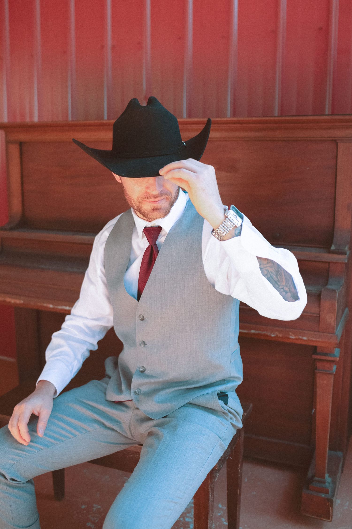 Groom in grey vest and black cowboy hat seated by vintage piano at western wedding.