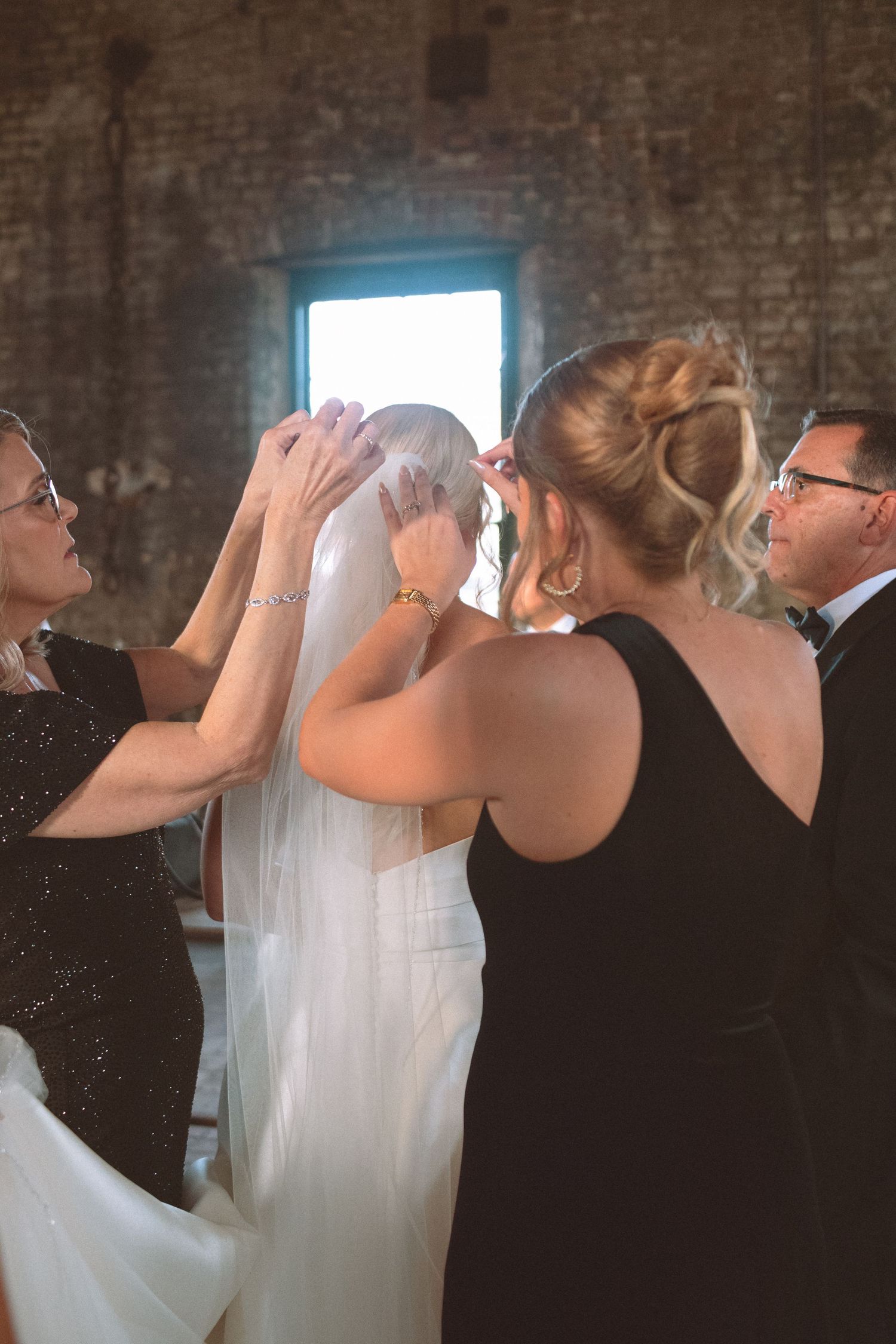 Bridesmaids and family members helping bride adjust her veil inside a rustic brick wedding venue.