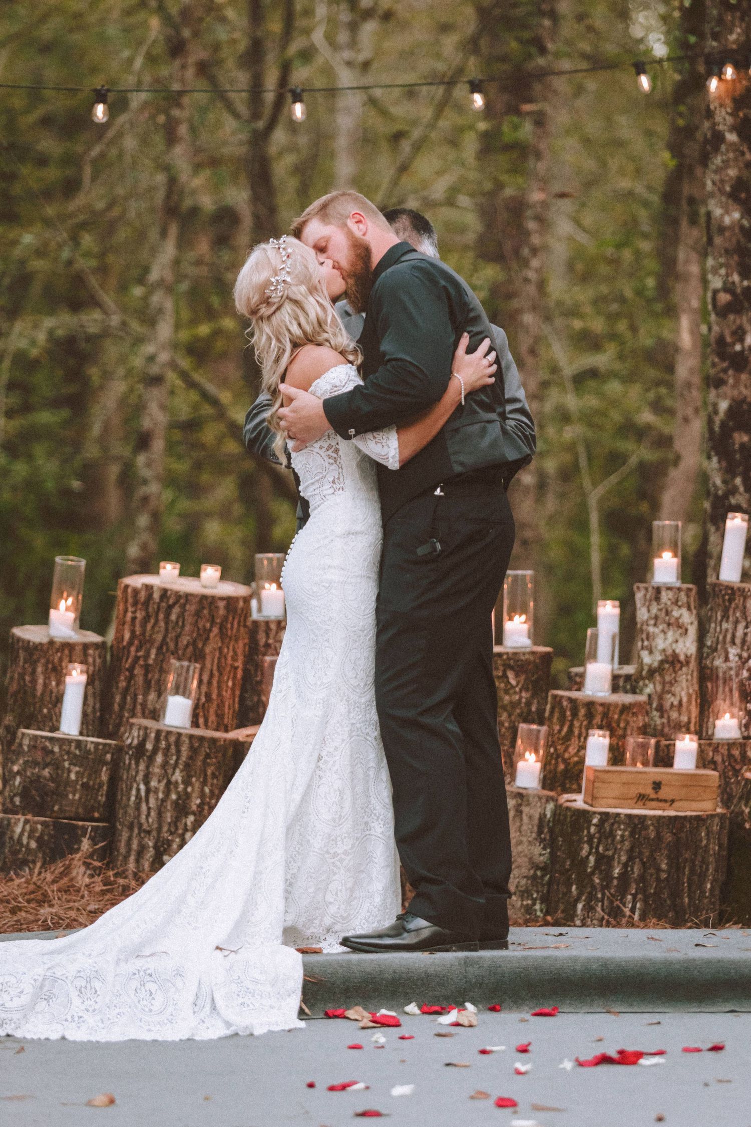 Bride and groom sharing their first kiss surrounded by candle-lit tree stumps at outdoor forest wedding.