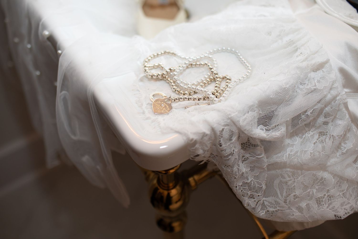 Pearl necklace with gold heart charm draped over white lace wedding dress on ornate gold table.