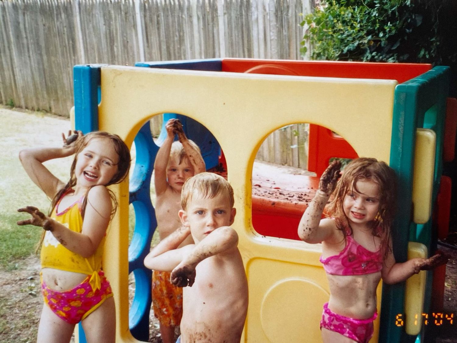 Four young children in swimsuits playing and posing in a colorful backyard plastic play structure.
