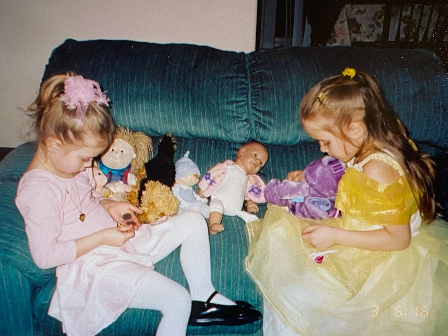 Two young girls in dress-up clothes play with dolls and stuffed animals on a blue sofa.