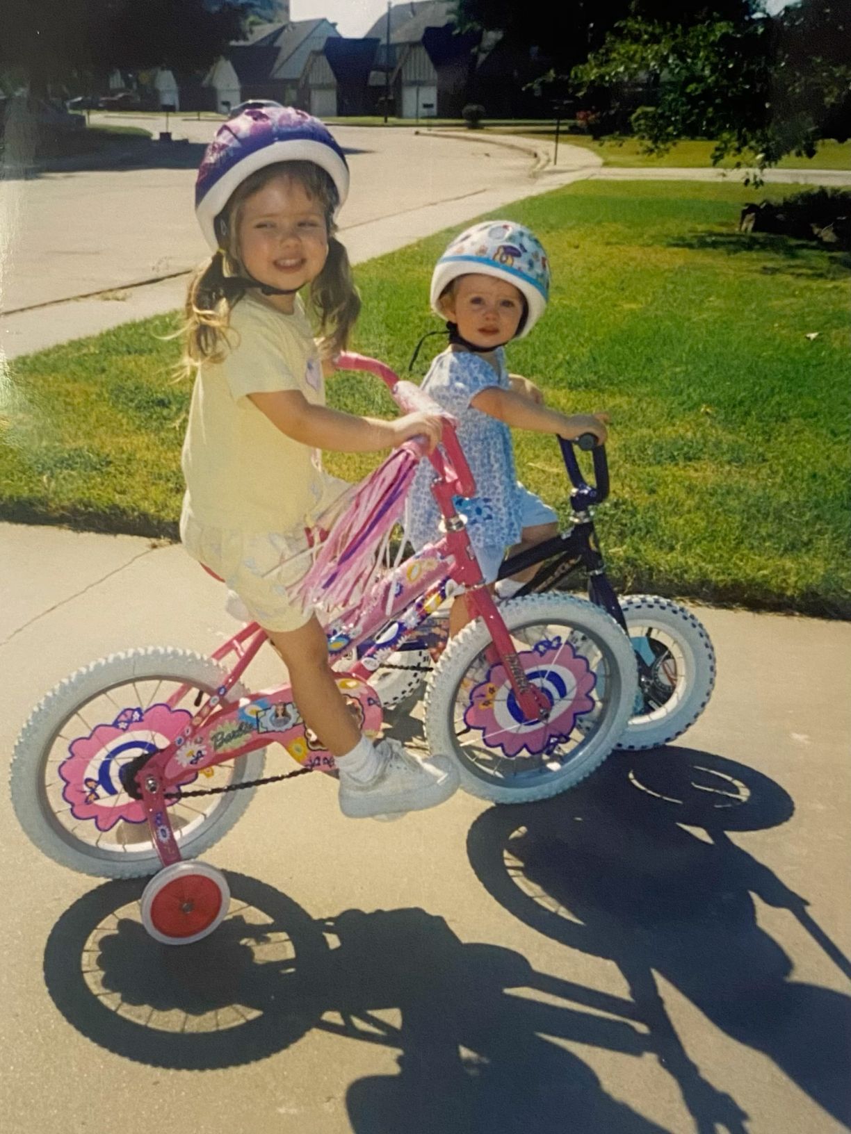 Two young girls wearing helmets riding bikes with training wheels on a sunny sidewalk.