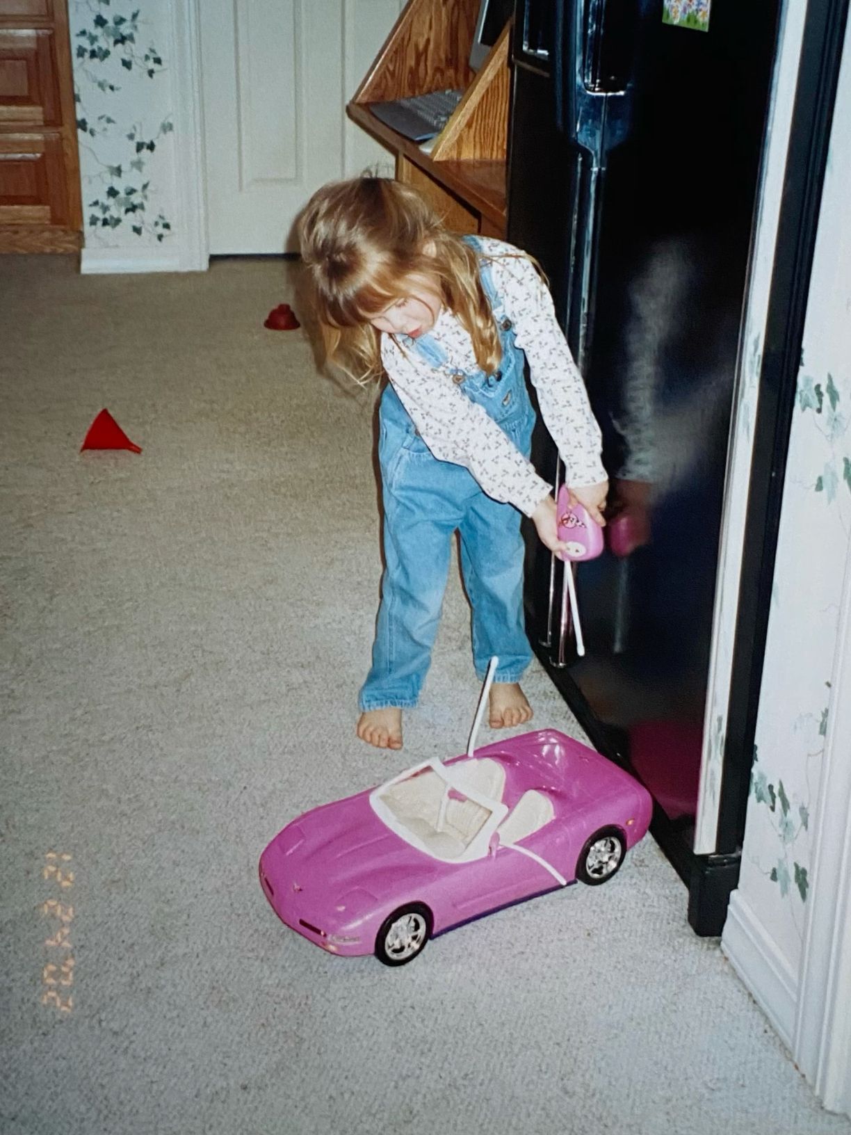 A young girl in denim overalls playing with a pink Barbie convertible car on a carpeted floor.