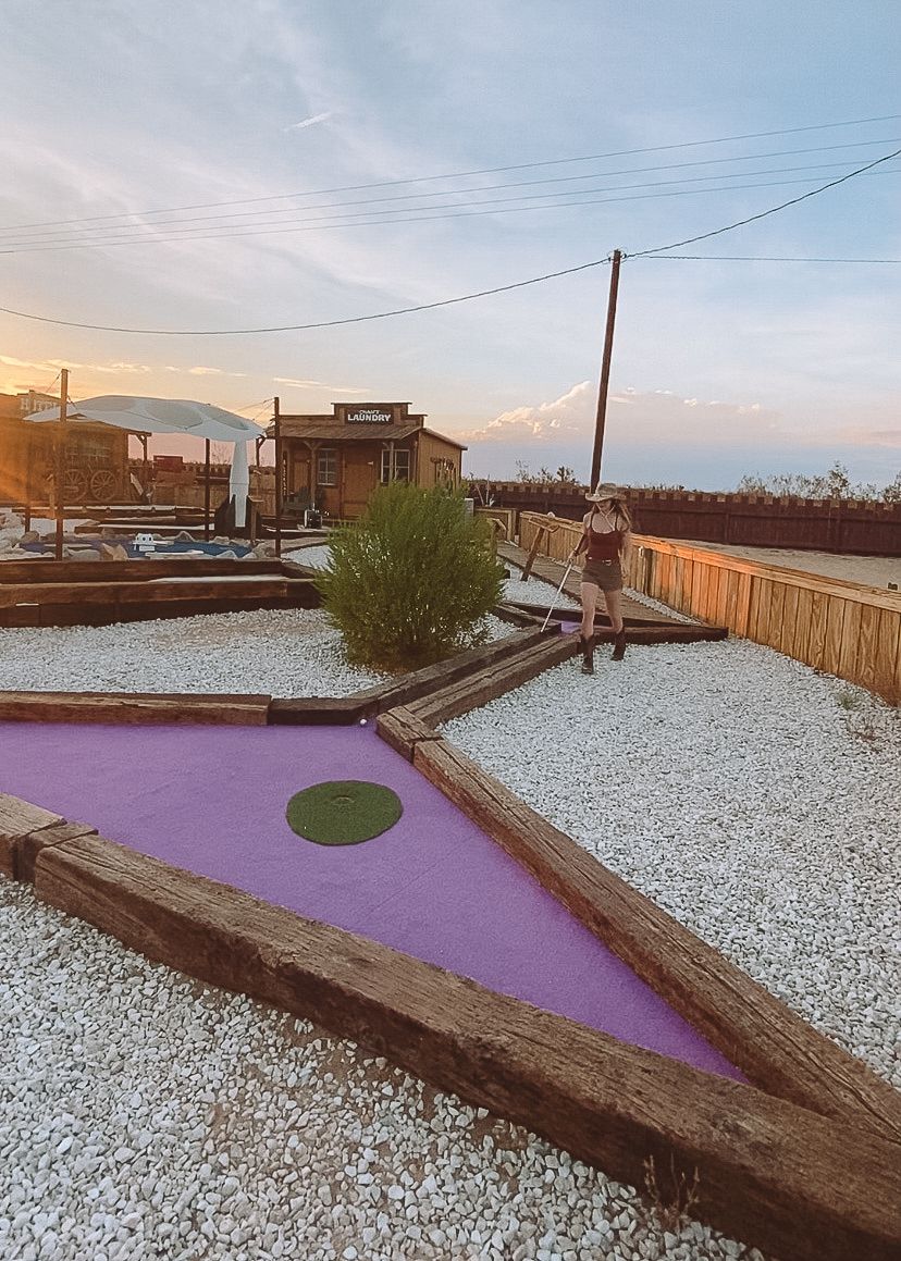 Woman playing mini golf at sunset with mountain views in background.