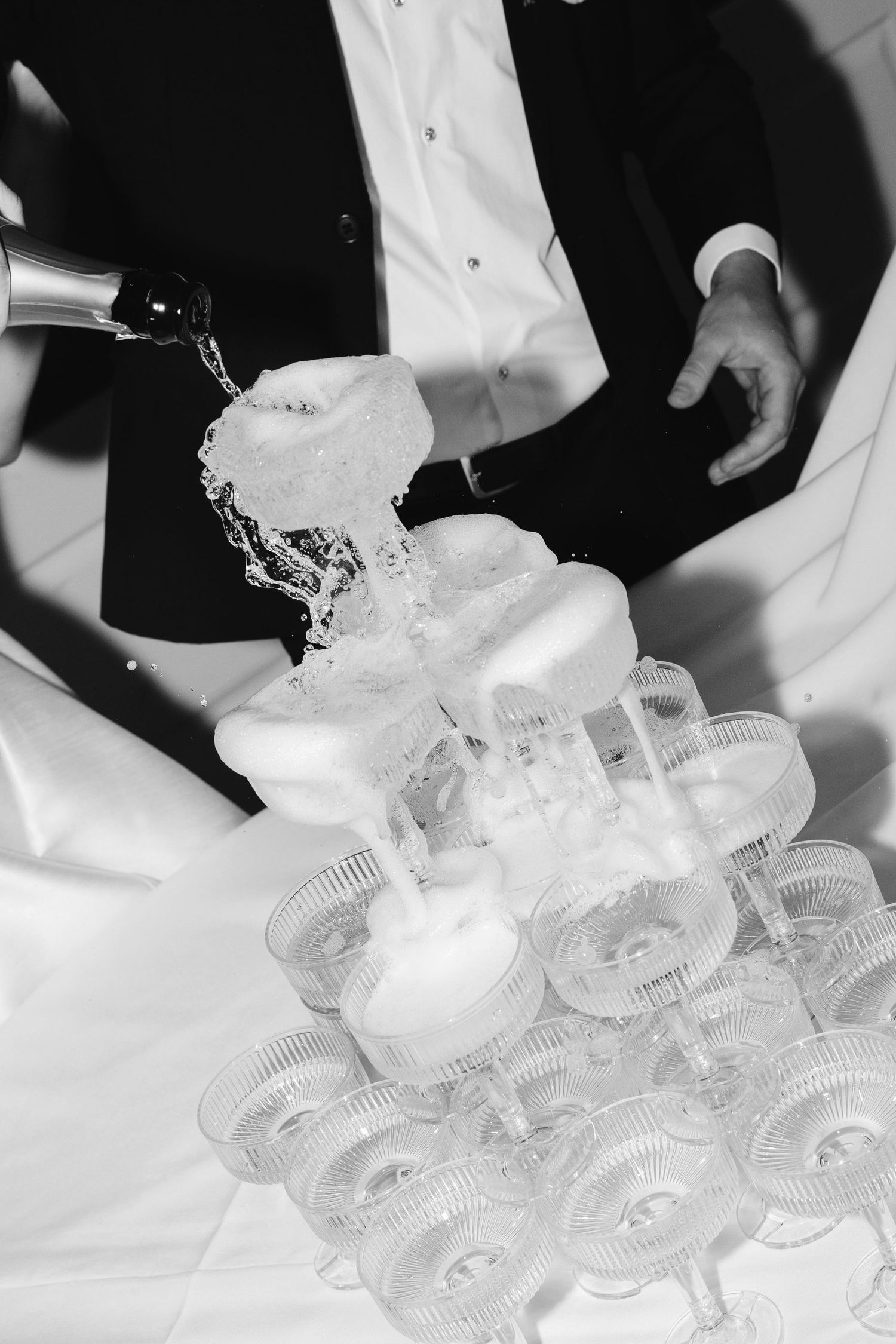 Black and white photo of a man in tuxedo pouring champagne into a tower of glasses at a formal event.
