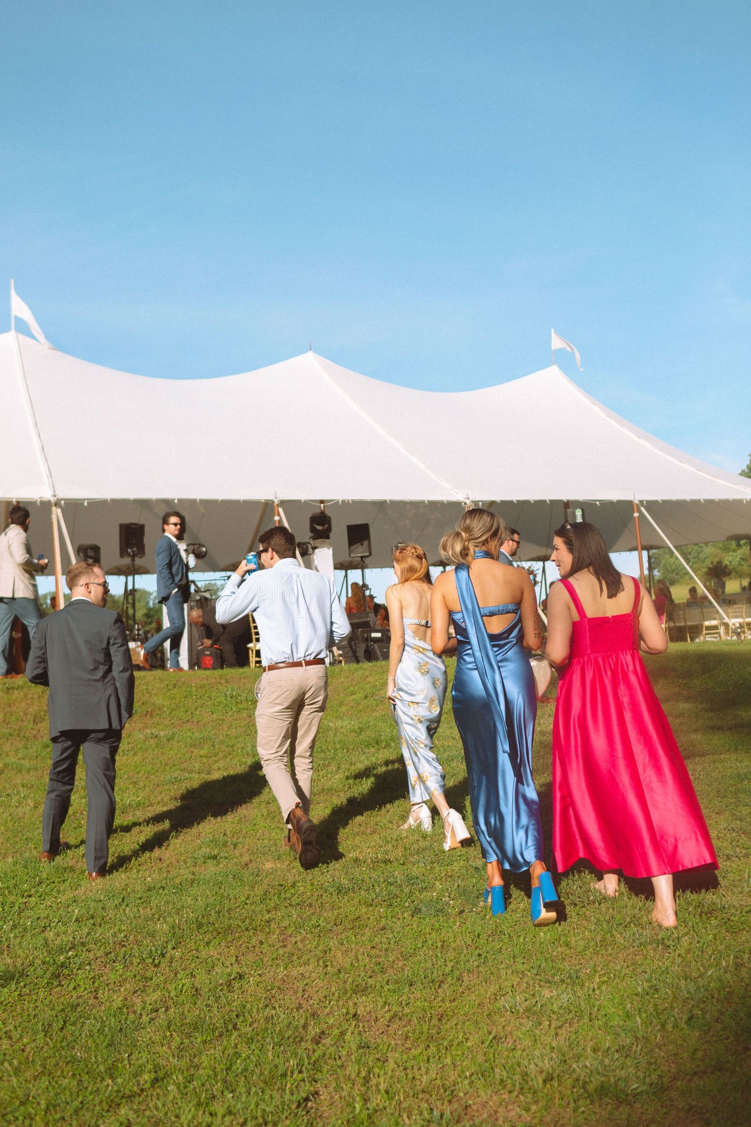Wedding guests walk toward large white tent reception venue on sunny green lawn.