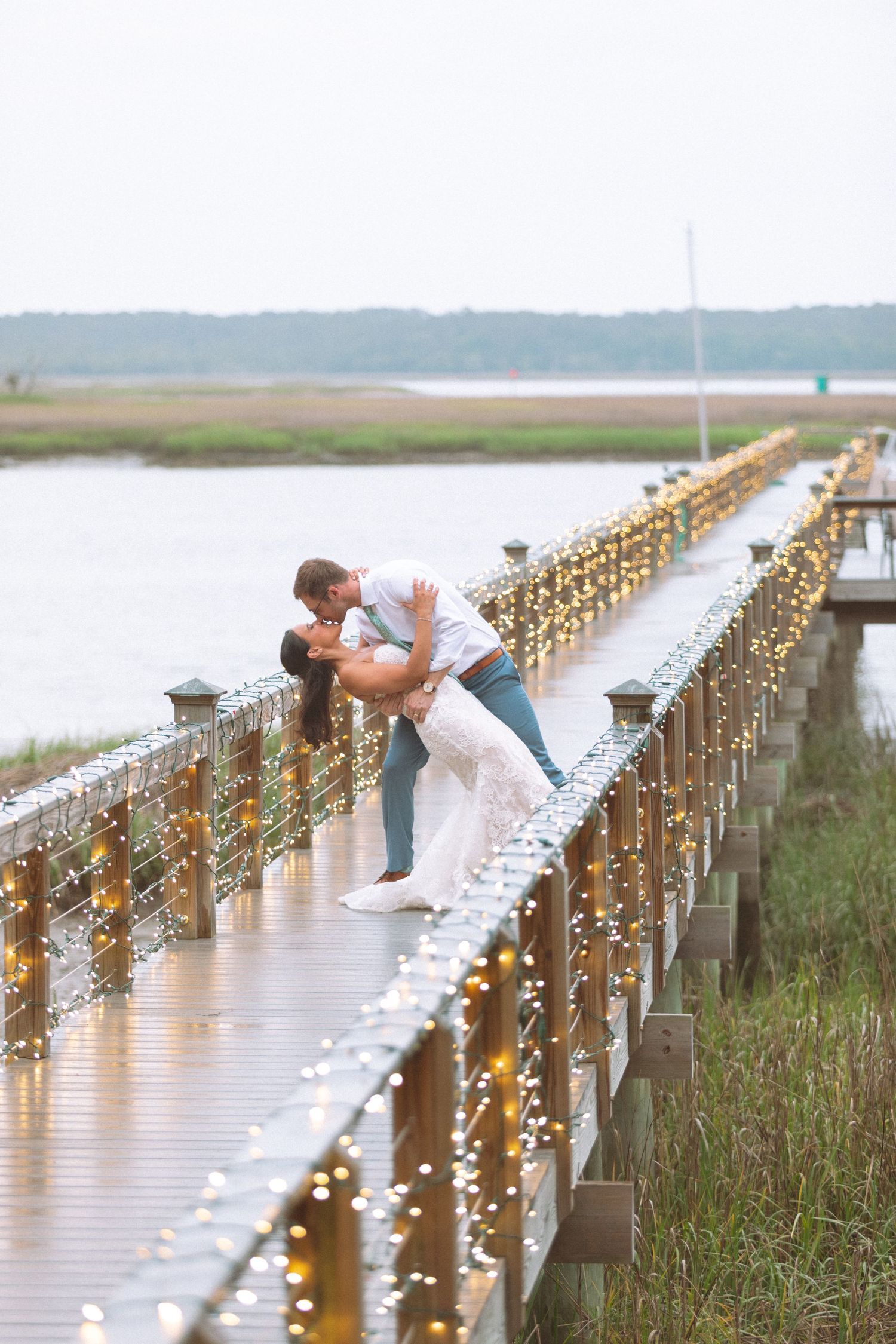 Couple shares romantic dip kiss on fairy light decorated wooden bridge over marsh at dusk.