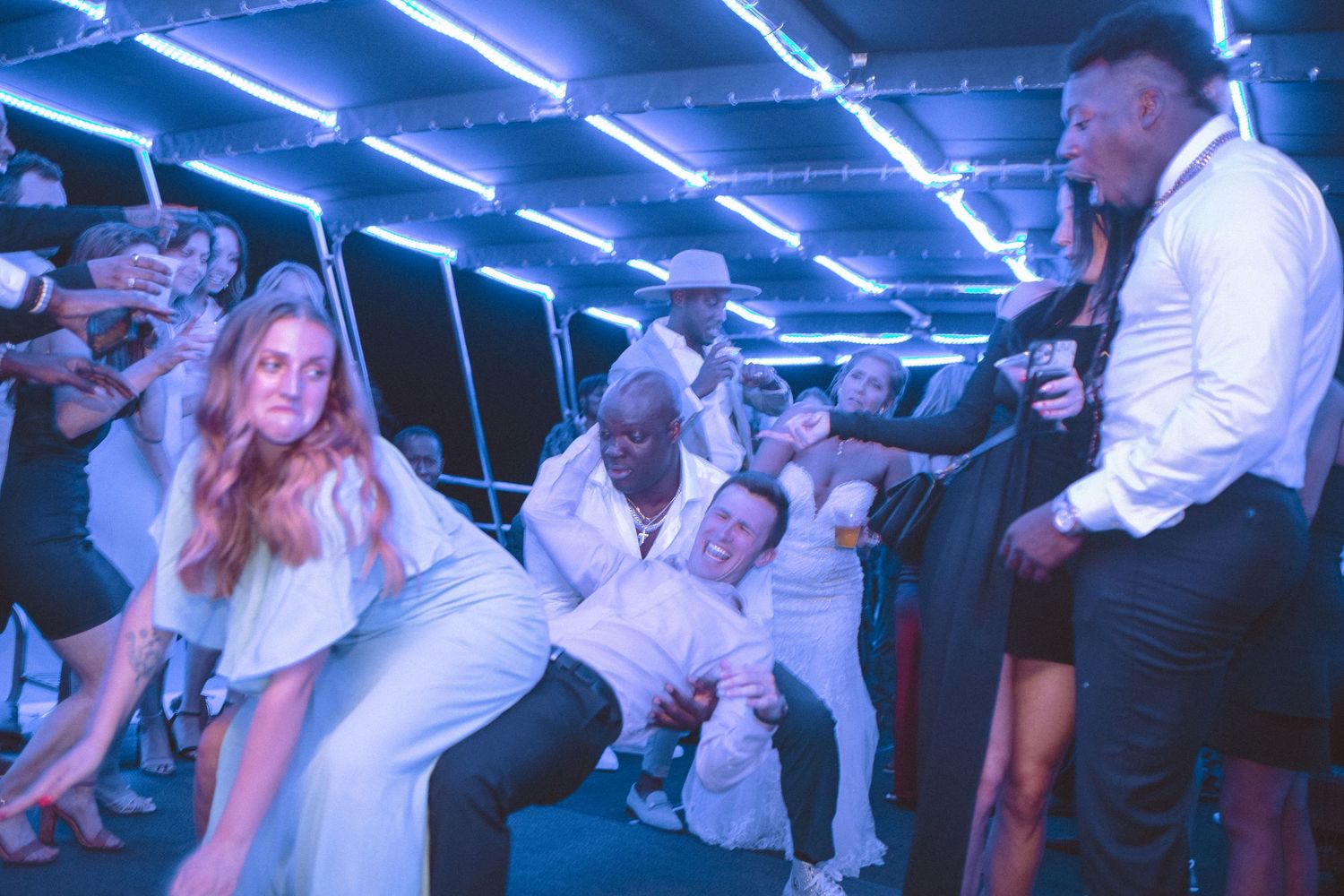 Energetic wedding reception dance floor with guests in white attire under blue neon lights.