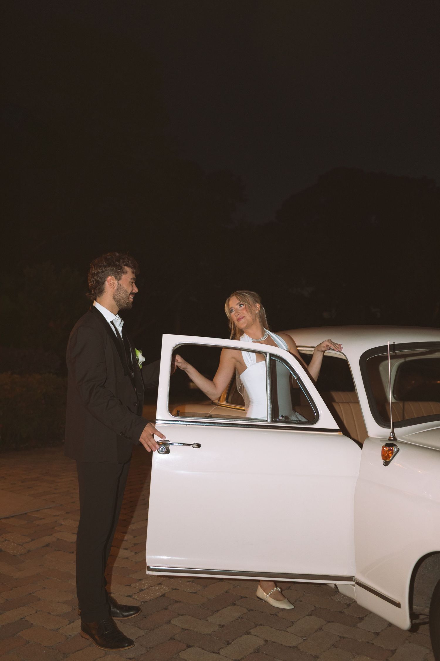 Groom opening door of vintage white car for bride outside at night.
