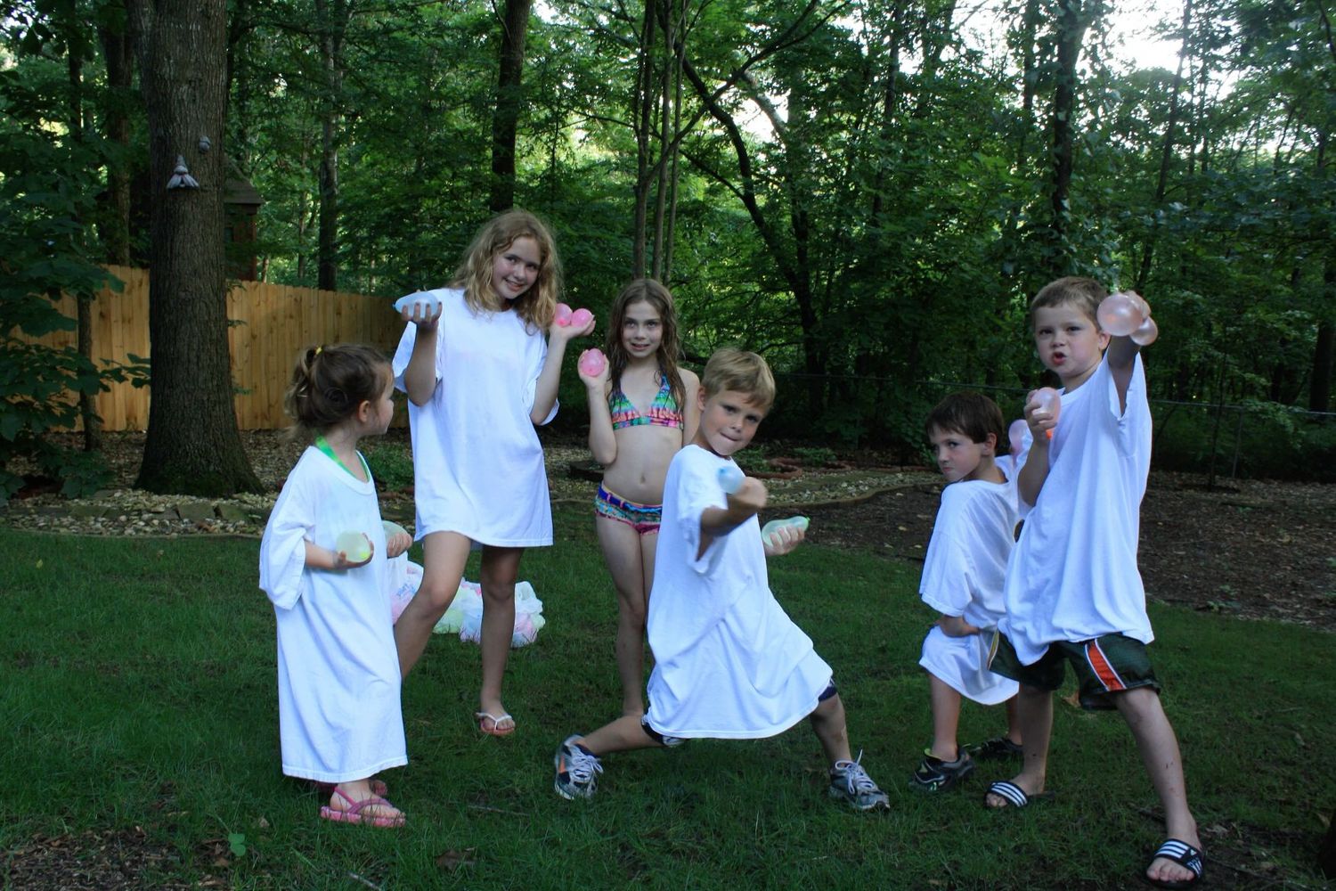 Group of children playing and posing in white t-shirts in a backyard surrounded by green trees.