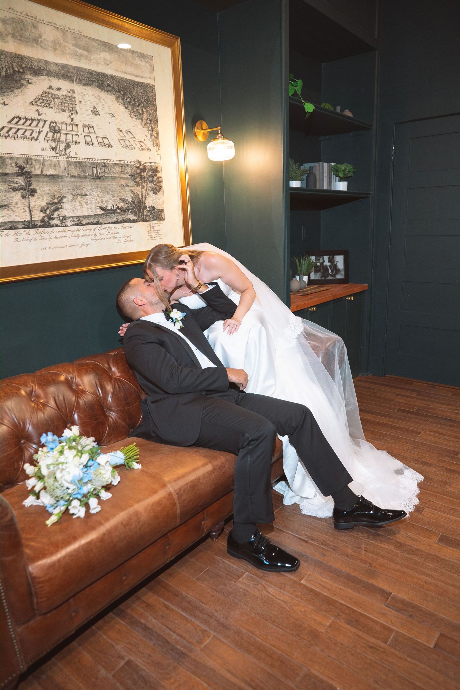 Groom sits on leather sofa as bride leans in for a kiss beside a blue bridal bouquet.